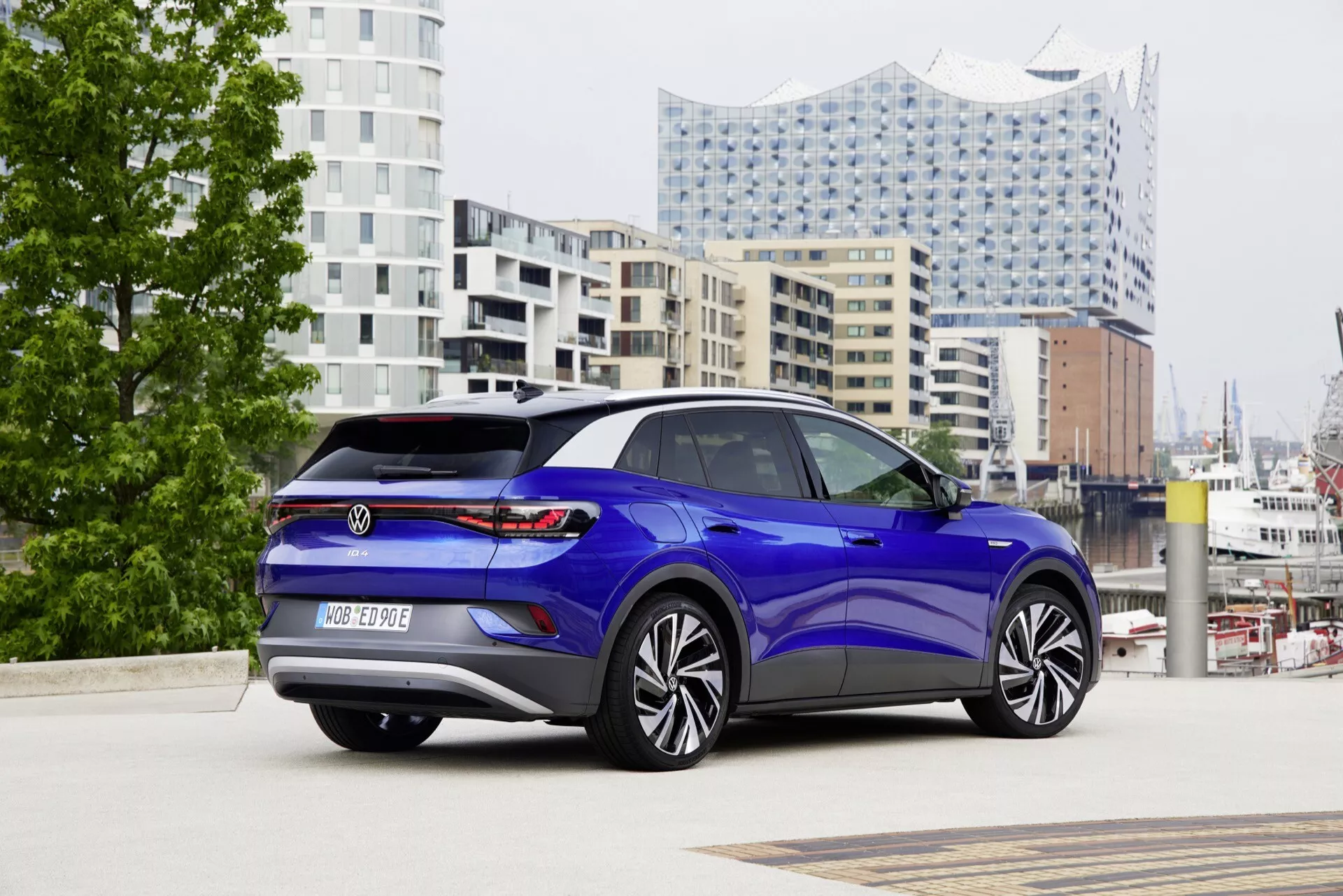 VW ID.4 1. generacji z 2020 roku, sfotografowany z perspektywy tyłu i boku, ukazujący cechy designu tylnej części, w tym LED-owe światła tylne, tylny zderzak z zintegrowanymi czujnikami parkowania i logo, a także aerodynamiczny profil boczny z dużymi kołami ze stopów lekkich wieloramienne, klamki drzwi w kolorze karoserii i przyciemniany dach szklany lub panoramiczny dach słoneczny, na tle miejskiego krajobrazu z nowoczesną architekturą.