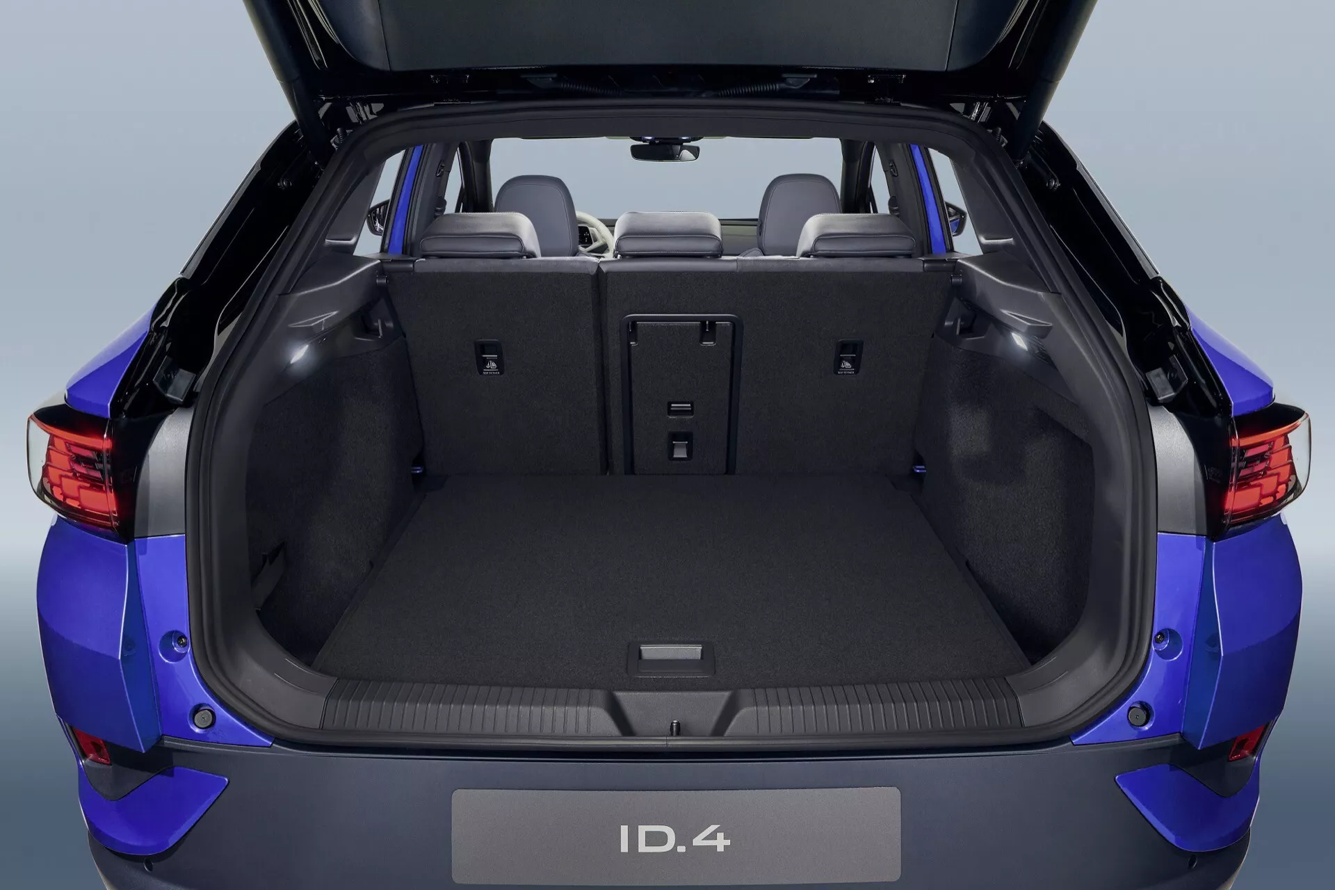 VW ID.4 2020 1. generacja: nowoczesne niebieskie elektryczne SUV widok z tyłu pokazujący otwarty luk bagażowy z przestronnym i uniwersalnym wnętrzem, złożone tylne siedzenia, bagażnik wykończony dywanikiem i zaawansowaną technologią czujników parkowania.