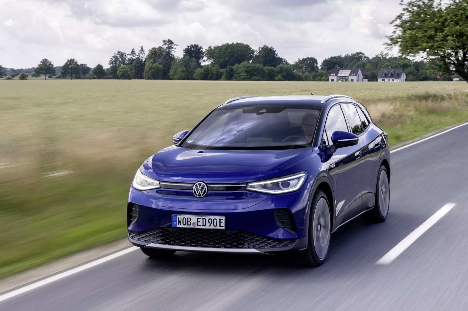 Niebieski elektryczny SUV Volkswagen ID.4 w ruchu, widziany z niskiego, przednio-bocznego kąta, podkreślający jego eleganckie światła LED, zamkniętą kratkę, felgi aluminiowe i dynamiczne linie designu na tle wiejskim.