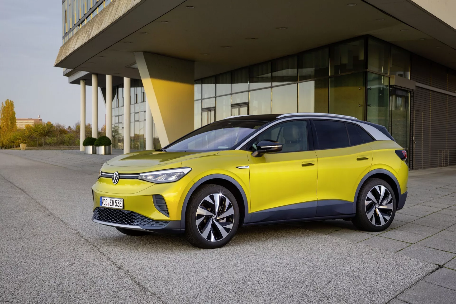 VW ID.4 1. Generacja 2020 w żywej żółci, z LEDowymi reflektorami, aerodynamicznymi kołami i eleganckim designem uchwyconym z perspektywy trzy czwarte frontalnej na tle nowoczesnej architektury, podkreślając jej elektryczne, efektywne i ekologiczne cechy w świetle dziennym
