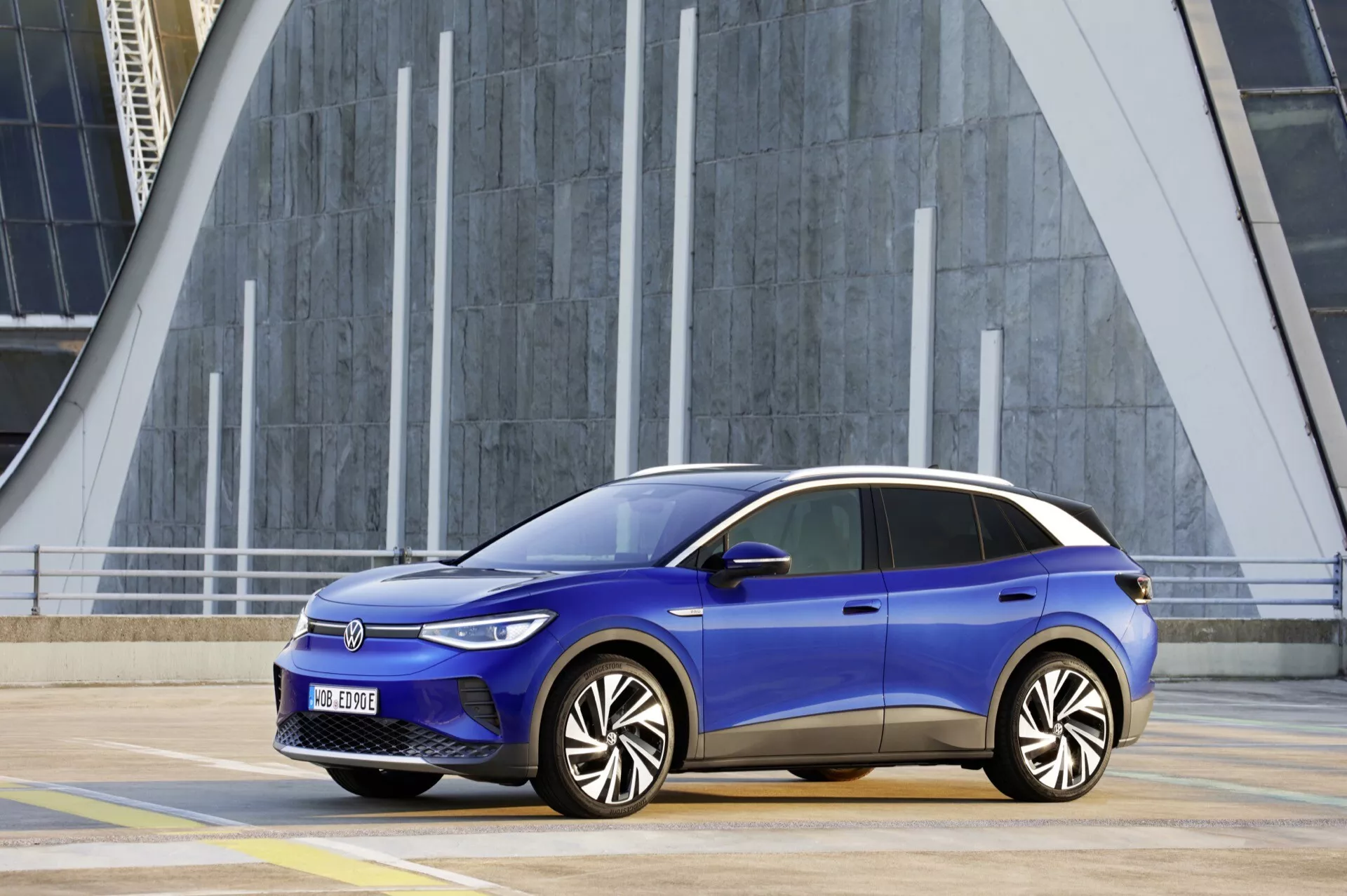 Boczny profil niebieskiego Volkswagena ID.4 2020 z aerodynamiczną konstrukcją, eleganckimi poziomymi reflektorami, wieloramienne kołami ze stopu i wskaźnikami LED na tle nowoczesnego miejskiego krajobrazu z geometrycznymi strukturami i pionowymi liniami