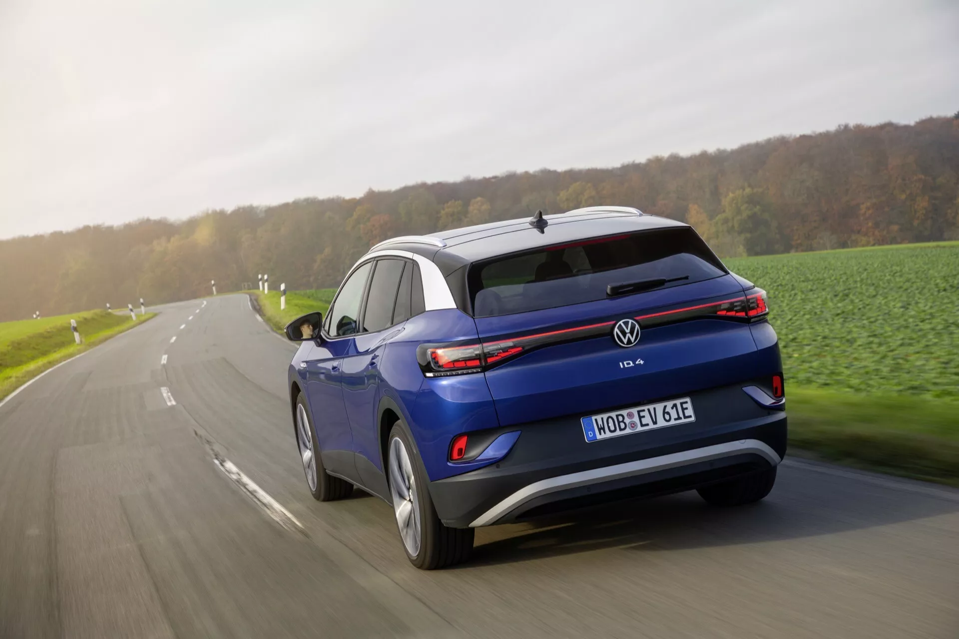 Dynamiczny widok z tyłu trzy czwarte niebieskiego elektrycznego SUV-a Volkswagen ID.4 w ruchu na utwardzonej drodze, przedstawiający diodowe światła tylne, spoiler dachowy, błyszczącą karoserię i widoczną tablicę rejestracyjną z oznaczeniem 'E', wskazującą na status pojazdu elektrycznego.
