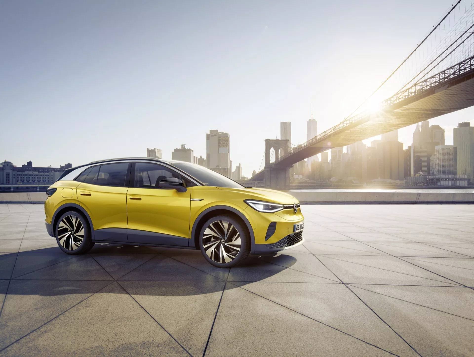 Hatchback/sedan VW ID.4 1 generacji 2020 w jaskrawo żółtym kolorze pod światłem złotej godziny, prezentujący profile przednie i boczne z ledowymi reflektorami i wieloramienne alufelgi, w miejskim otoczeniu z tłem mostu