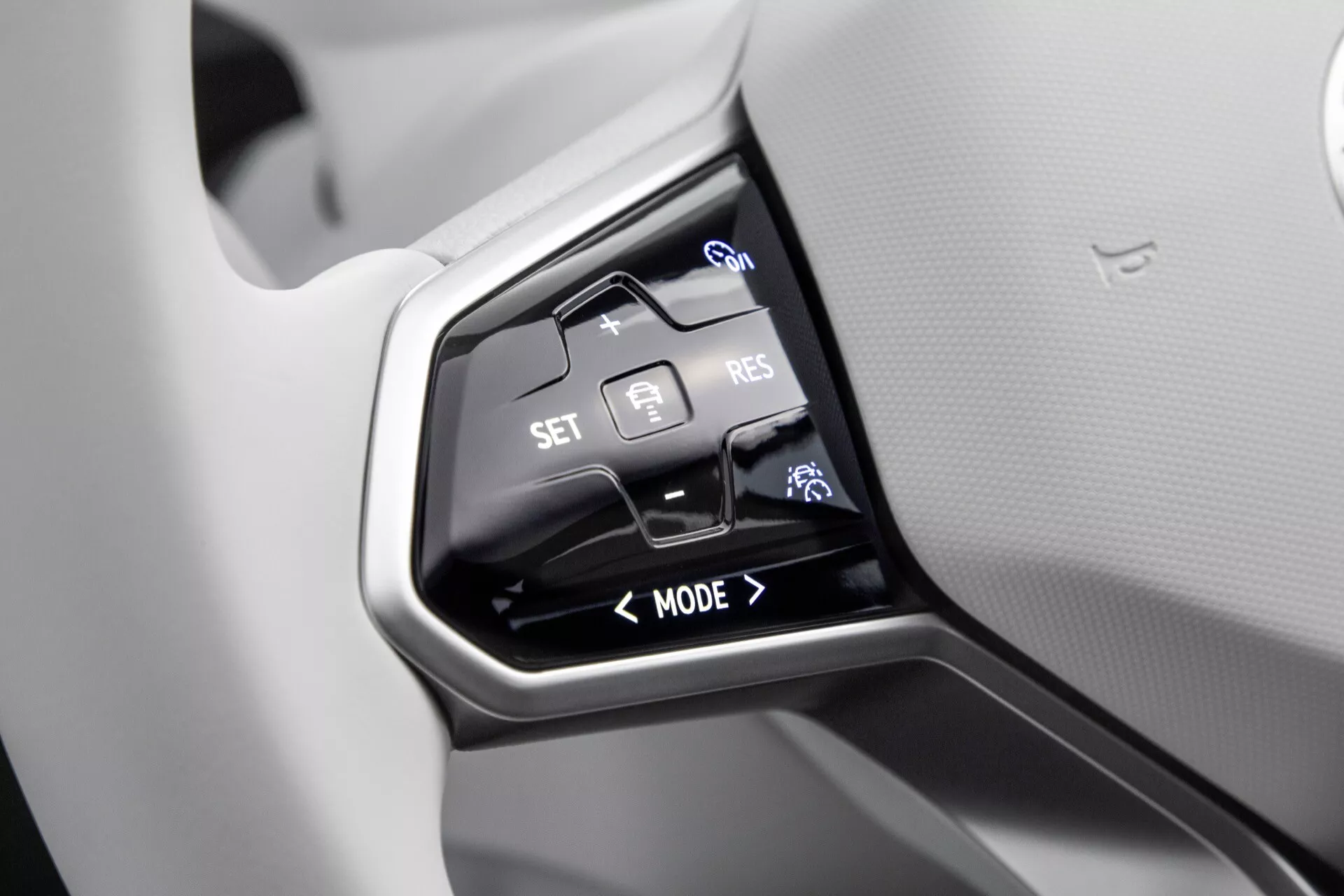 Zbliżenie na przyciski kierownicy VW ID4 1. generacji 2020 skupiające się na adaptacyjnym tempomacie, trybach jazdy i funkcjach sterowania głosem, umieszczone na błyszczącym czarnym panelu na tle jasnoszarej kierownicy.