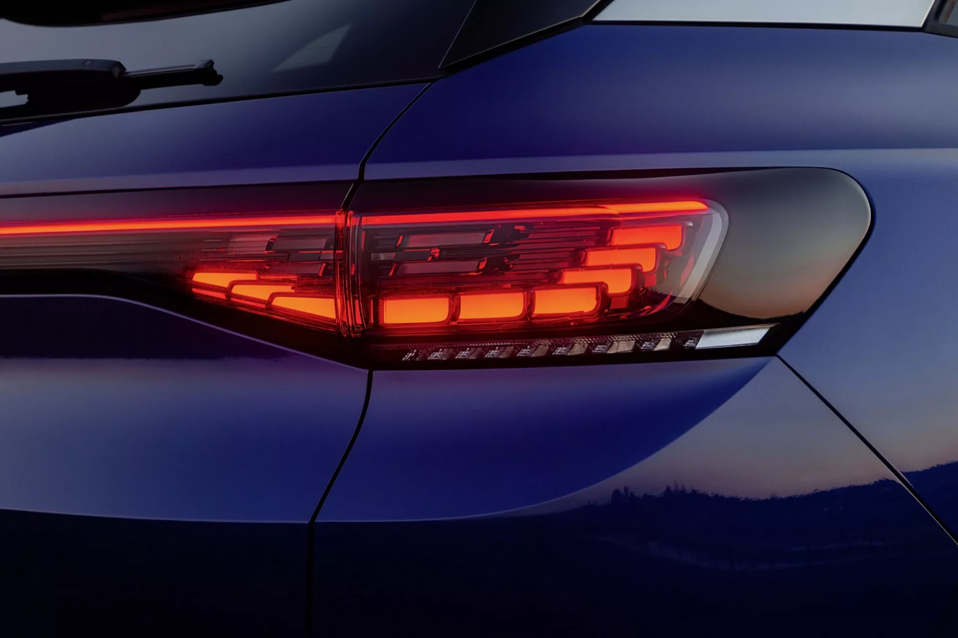 Zbliżenie na nowoczesną konstrukcję tylnej lampy LED VW ID.4 1. generacji z 2020 roku, prezentując jej smukły poziomy pasek i złożone oświetlone segmenty na tle odblaskowej głębokiej niebieskiej karoserii