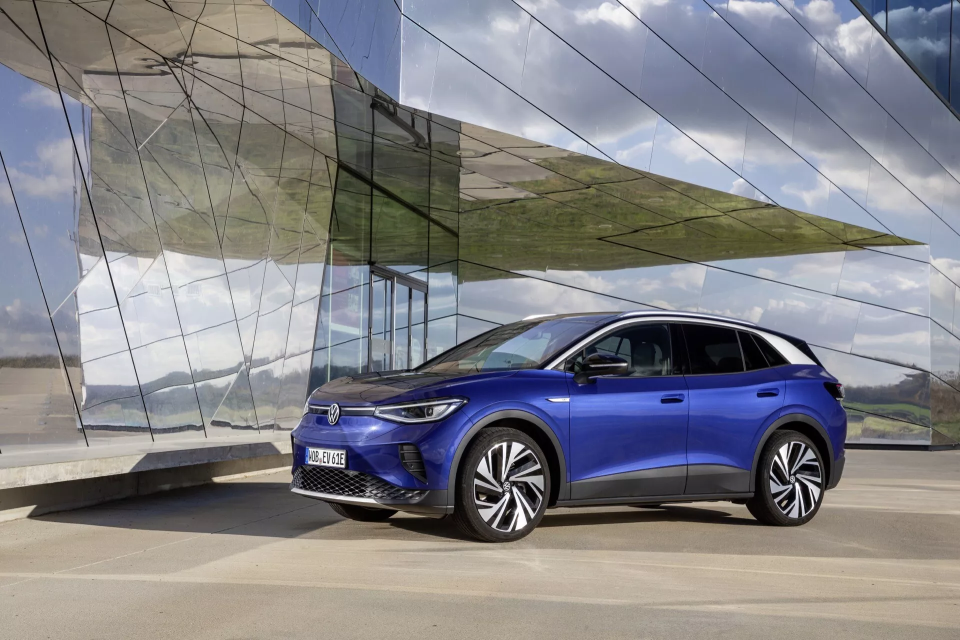 VW ID.4 1. generacja 2020 w żywym niebieskim, prezentujący nowoczesny design SUV z widoku trzy czwarte z przodu na gładkim betonie, na tle architektury kątowej ze szkła i metalu, wyświetlający reflektory LED, unikalne koła ze stopu i elegancki profil boczny.