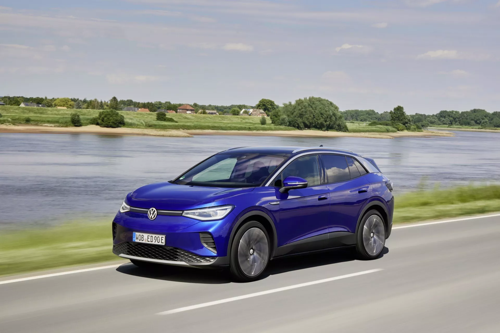 Dynamiczny trzykwartalny widok z przodu na elektryczny samochód VW ID.4 2020 w jaskrawo niebieskim kolorze, prezentujący jego elegancki design i ruch na tle naturalnej autostrady z spokojnym akwenem i zielenią