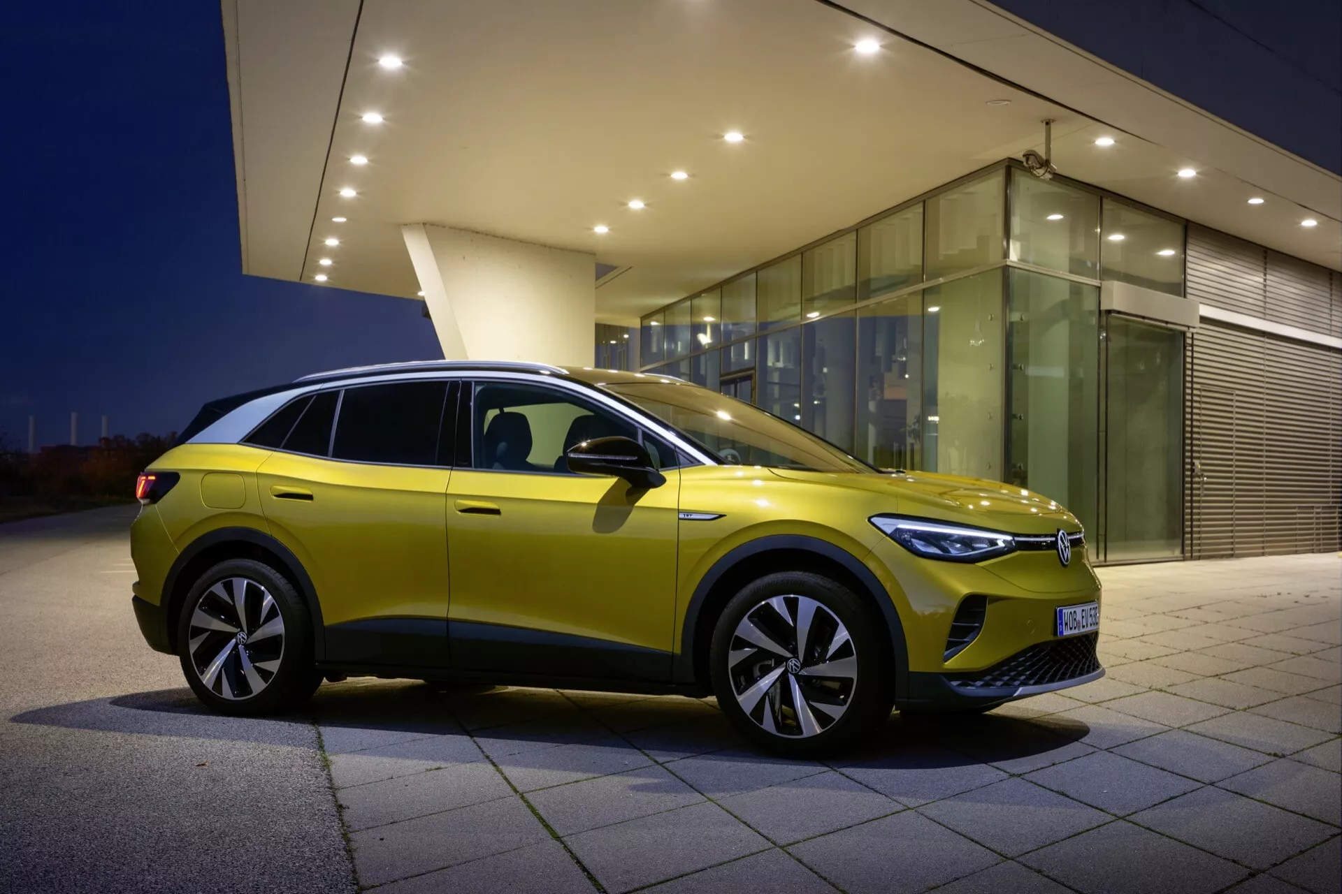VW ID.4 1. generacji rok 2020 w żywym żółtym kolorze, z widoku trzech czwartych z przodu, podkreślający jego diody LED, masywną karoserię i duże felgi aluminiowe na tle miejskim o zmierzchu
