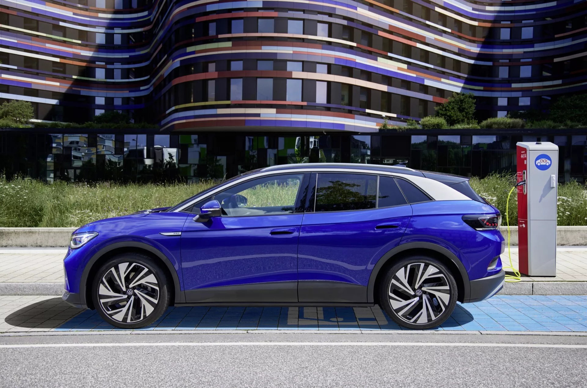 Widok boczny niebieskiego pojazdu elektrycznego VW ID.4 z 2020 roku, podkreślający jego boczne elementy projektowe, od eleganckich przednich świateł LED i zamkniętej kraty, poprzez klamki drzwiowe w linii z karoserią i konturowane linie nadwozia, po panoramiczny dach szklany i koła ze stopów lekkich, z samochodem podłączonym do stacji ładowania.