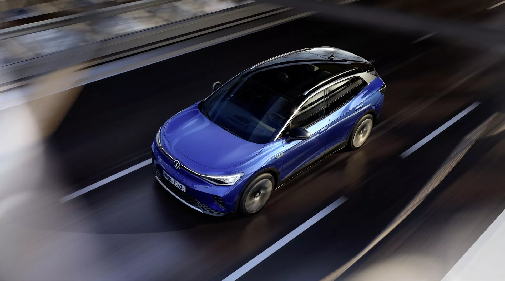 Dynamiczny widok z góry na nowoczesne niebieskie VW ID.4 2020 w ruchu, podkreślający jego elegancki design, szyberdach, konturowany kaptur i aerodynamiczne nadwozie na tle rozmytej drogi, sugerujący zaawansowane funkcje i ekologiczną technologię w świetle dziennym.