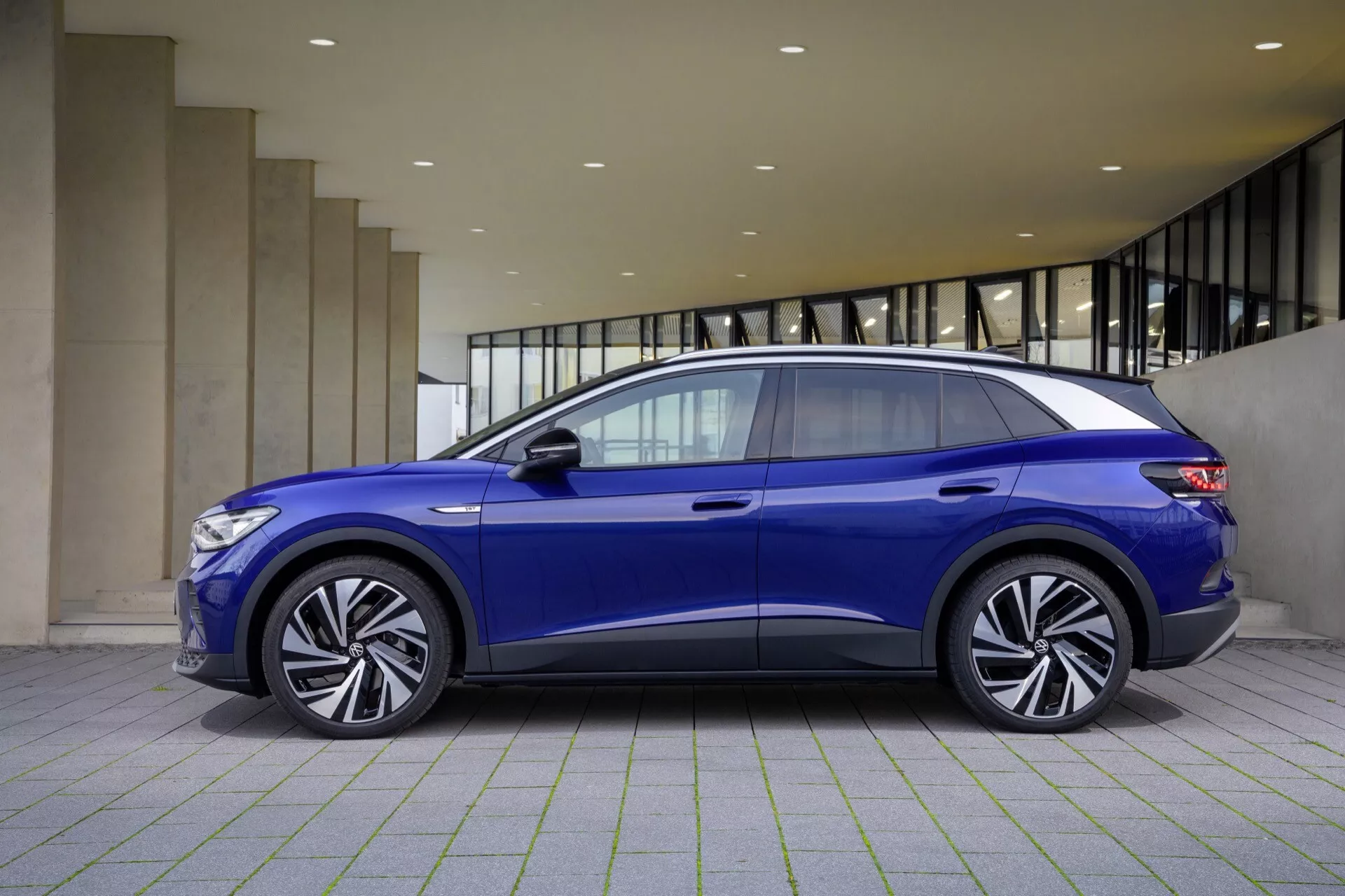VW ID.4 1. generacja 2020 w żywym niebieskim kolorze prezentujący swój aerodynamiczny profil boczny z opadającą linią dachu, charakterystyczną linią boczną nadwozia oraz zaawansowane funkcje technologiczne takie jak płaskie klamki, zintegrowane kierunkowskazy w lusterkach bocznych, duże koła z lekkich stopów wieloszprychowe i panoramiczny dach słoneczny, zaparkowany w nowoczesnym otoczeniu.