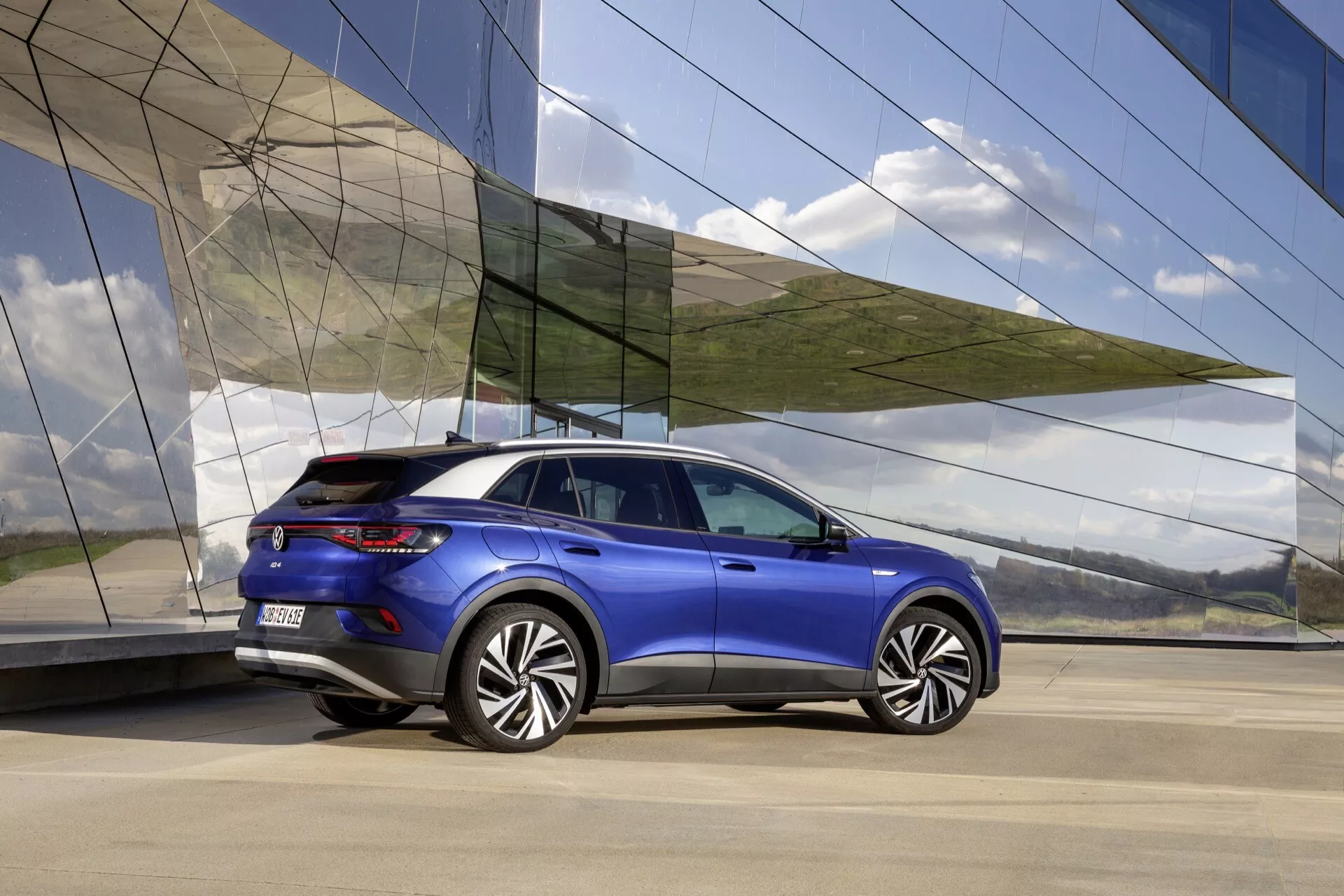 VW ID.4 1. Generacji 2020 w Metalicznym Niebieskim - Widok z Tyłu i Boku podkreślający Aerodynamiczny Design, Sportową Sylwetkę, Duże Koła z Stopów, Nowoczesne Światła LED i Premium Wykończenie na Tle Architektury