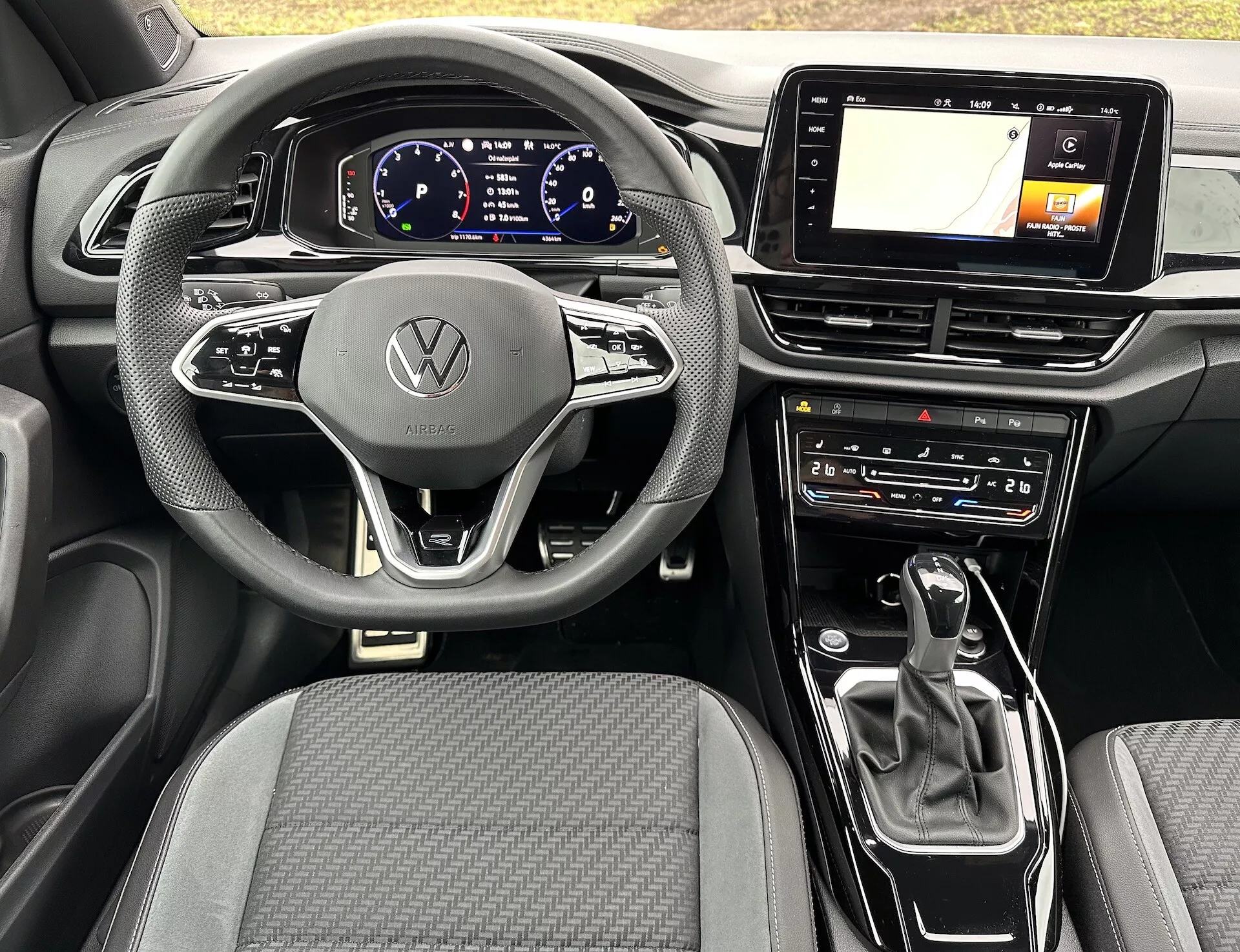 Widok wnętrza VW T-Roc Cabriolet 2022 z nowoczesną, trójramienną kierownicą z emblematem Volkswagen, cyfrowym zegarem, ekranem dotykowym systemu infotainment z GPS, selektorem biegów automatycznej skrzyni biegów oraz ustawieniami klimatyzacji, podkreślający zaawansowaną technologię i elegancki design auta