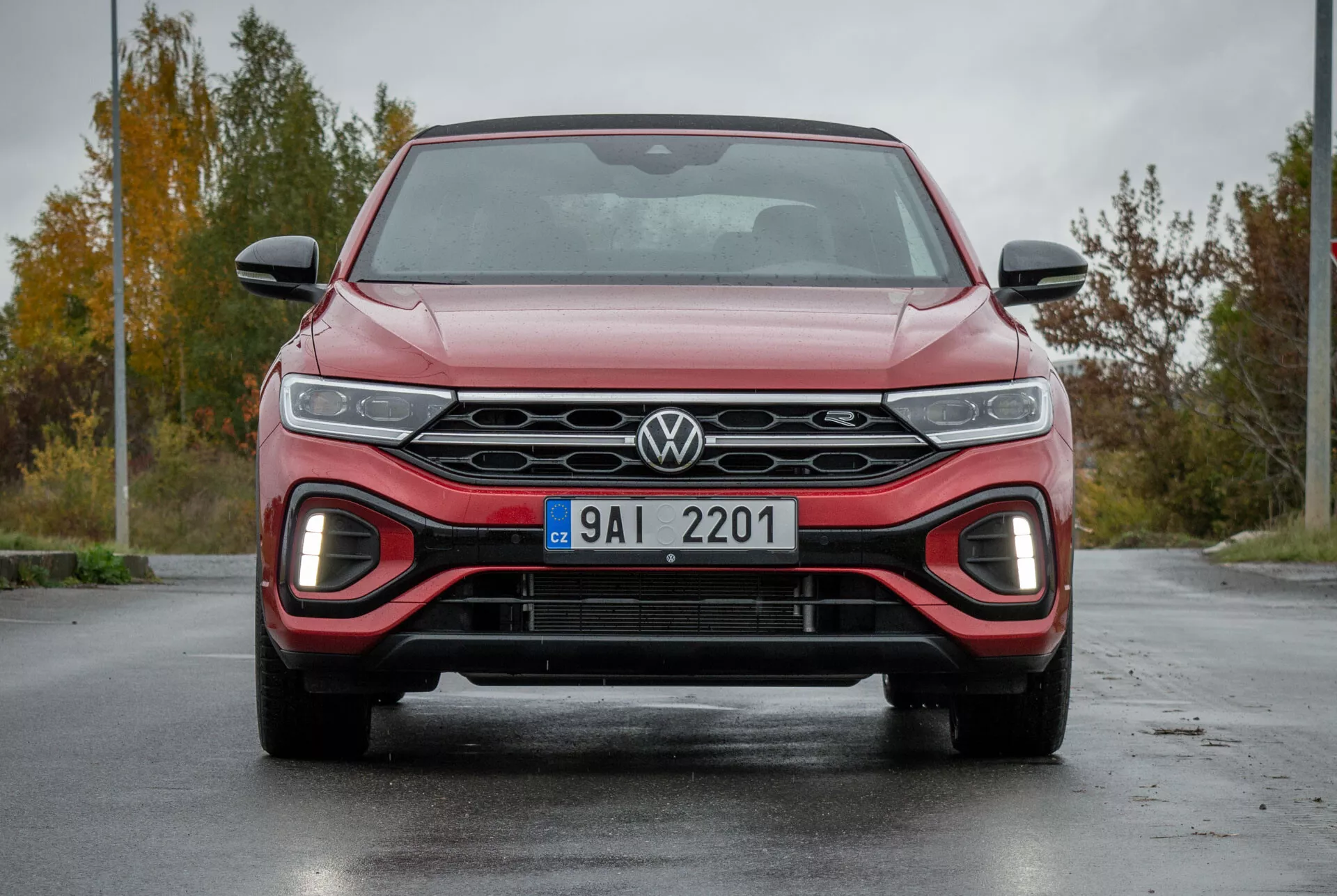 Widok z przodu VW T-Roc Cabriolet 2022 podkreślający elegancką kratkę z logo Volkswagen, reflektory LED, dolny wlot powietrza z czerwonym akcentem dla sportowego wyglądu i aerodynamiczny design na mokrej nawierzchni