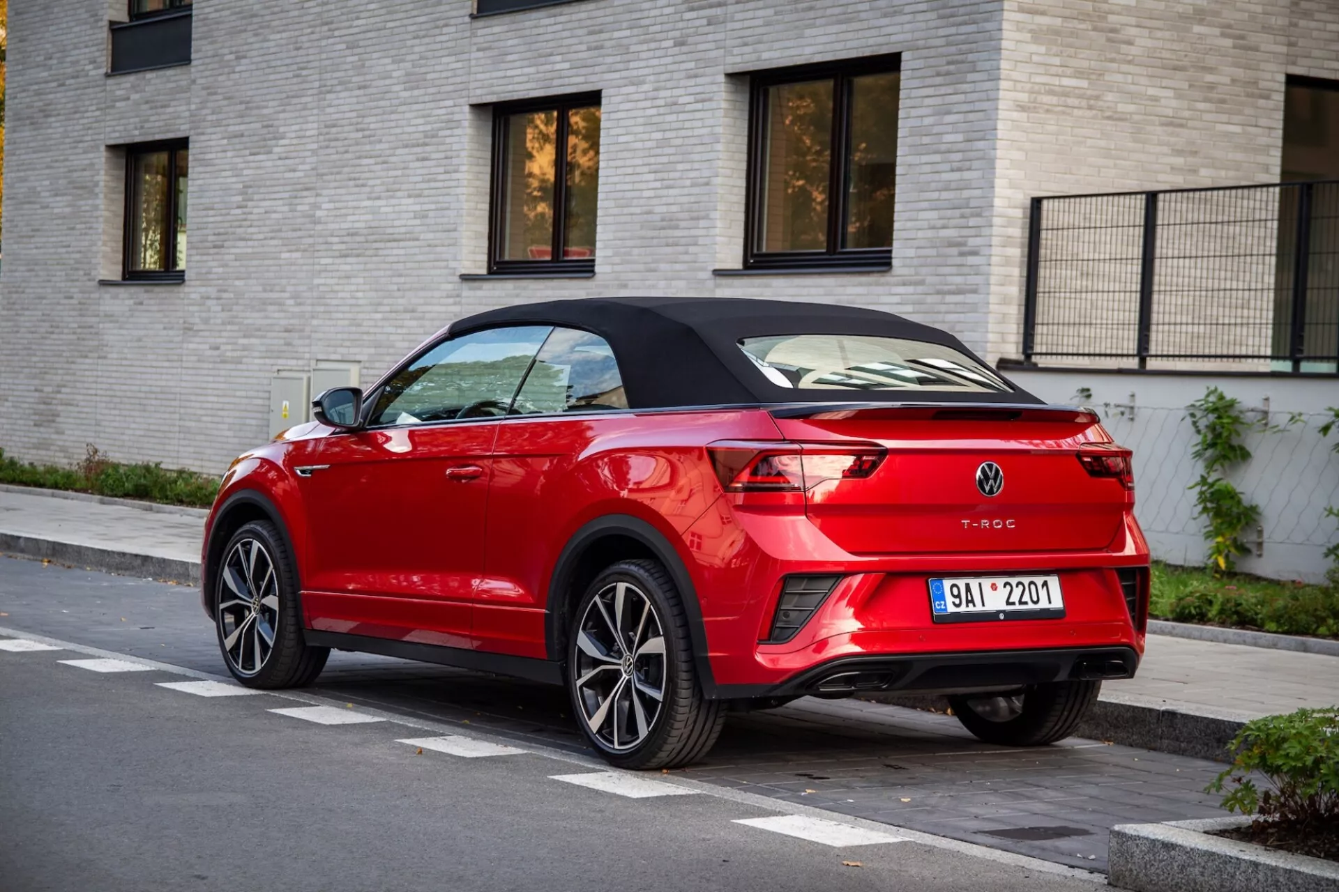 Tylny widok z trzech czwartych czerwonego kabrioletu Volkswagen T-Roc zaparkowanego na ulicy, podkreślający jego czarny składany dach, eleganckie tylne światła LED, tylny spojler, podwójne końcówki wydechu i zintegrowane czujniki parkowania na tle jasnoczerwonego budynku z cegły.