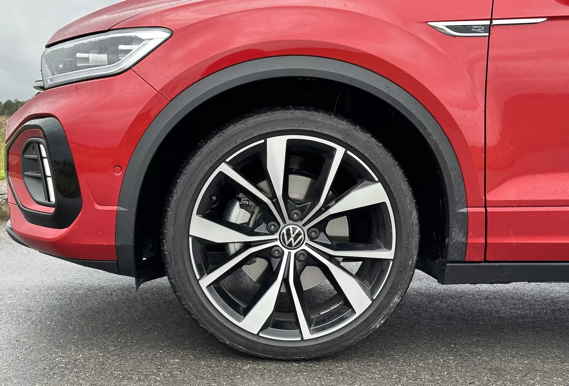 Zbliżenie na VW T-Roc 2022 skupiające się na wieloramienne koło ze stopu z dwukolorowym wykończeniem, oponę niskoprofilową i częściowo przedni błotnik prezentujący sportową czerwoną karoserię oraz detal oświetlenia LED