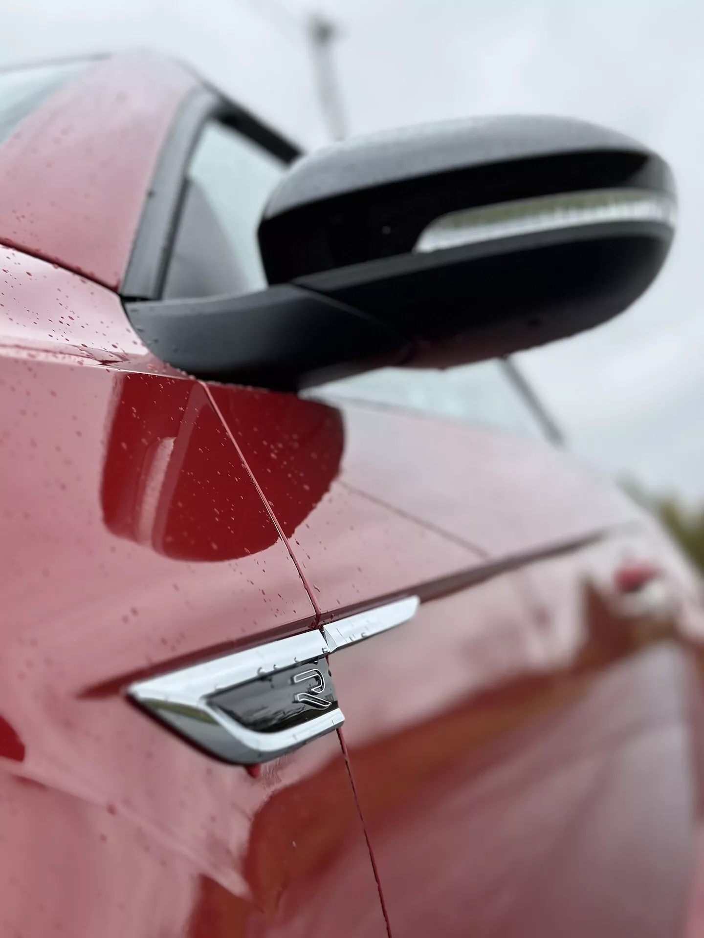 Zbliżony boczny widok na czerwone VW T-Roc Cabriolet 2022, podkreślający tylną część z detalami czarnego lusterka bocznego, klamki drzwi z systemem wejścia bezkluczykowego i elementów stylistycznych samochodu, w tym znaczku, pod lekko uniesionym kątem z kroplami wody na powierzchni wskazującymi na niedawny deszcz lub mycie, na rozmytym tle