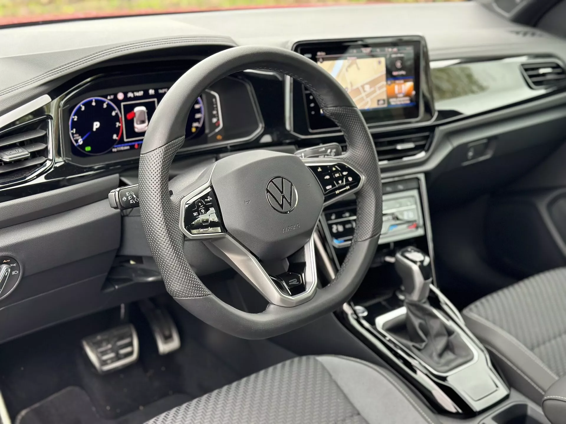 Wnętrze Volkswagen T-Roc Cabriolet 2022 z perspektywy kierowcy, na pierwszym planie trójramienna kierownica z multifunkcyjnymi przyciskami i logo Volkswagen, cyfrowy kokpit, system infotainment z nawigacją, kontrola klimatu, elegancka dźwignia zmiany biegów, sterowanie pomocą przy prowadzeniu, oraz premium design kokpitu z metalicznymi akcentami i fotelami z materiału przypominającego skórę.
