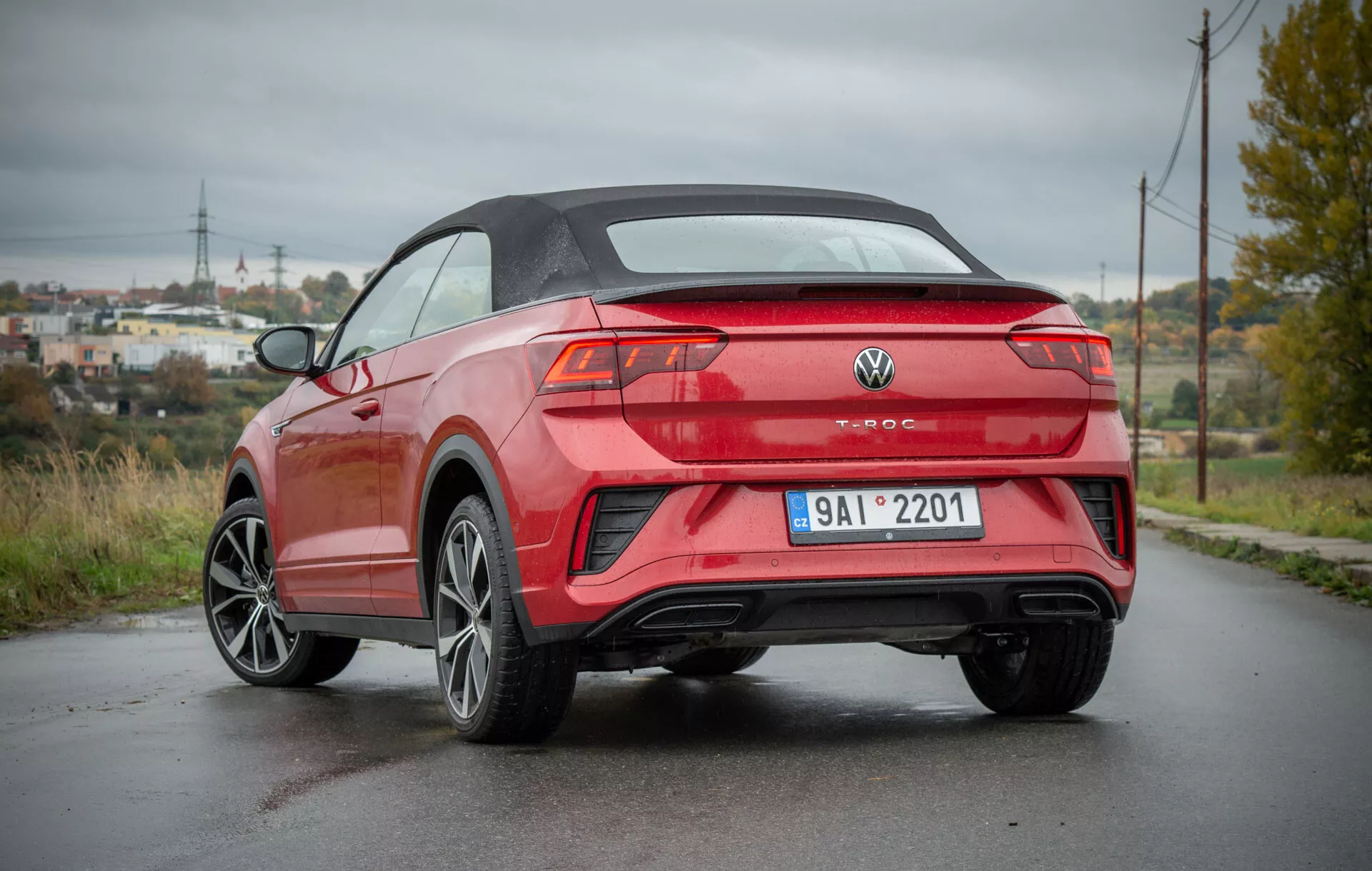 Tylny widok z trzech kwartałów VW T-Roc Cabriolet 2022 zaparkowanego na mokrej drodze, prezentujący jego nowoczesny design kabrioletu, ostre linie nadwozia, sportowe alufelgi, eleganckie tylne światła i podwójny wydech, na tle półwiejskiego krajobrazu z słupami elektrycznymi i bujną zielenią.