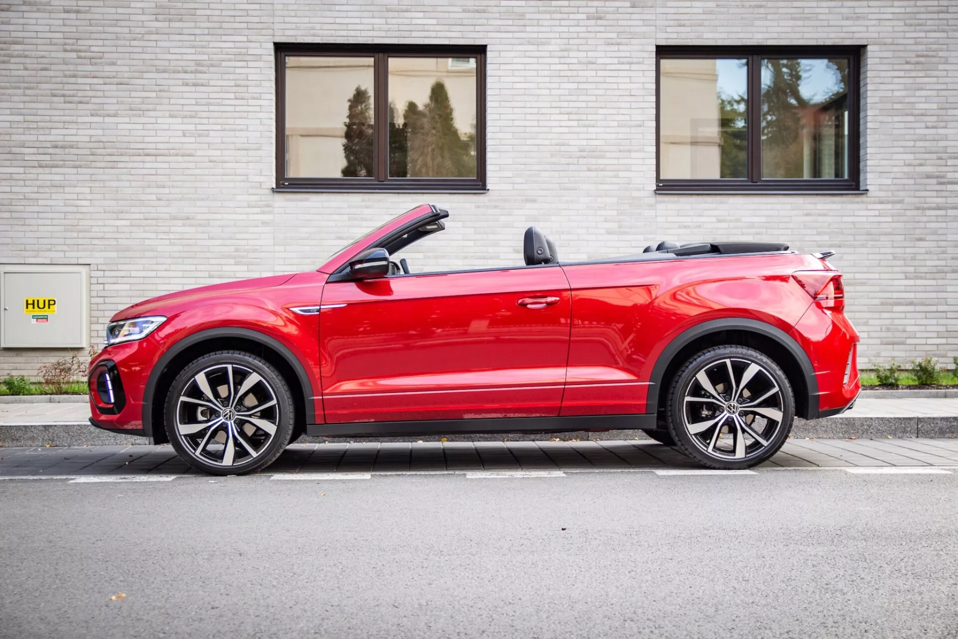 Czerwony kabriolet Volkswagen T-Roc 2022 zaparkowany równolegle do szarej ściany z cegieł z napisem HUP, pokazujący profil boczny z lekkim kątem od przodu, składany dach, światła LED i sportowe felgi aluminiowe