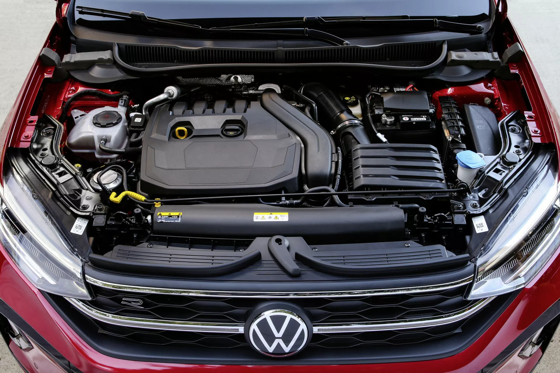 Zbliżony widok na komorę silnika Volkswagen Taigo 2021 z otwartą maską, pokazujący komponenty silnika, logo Volkswagen na grillu oraz szczegółowy projekt inżynierski