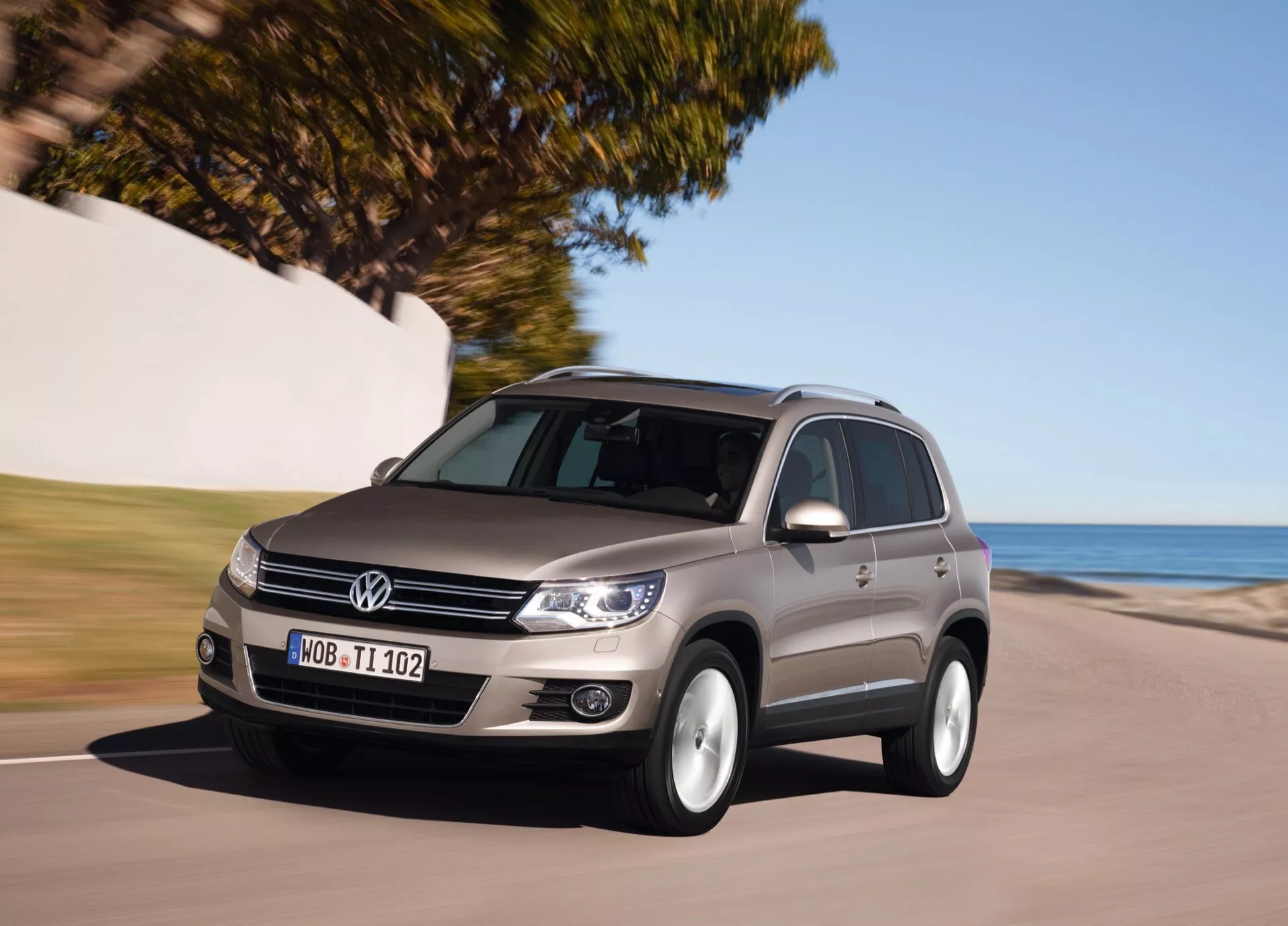 SUV średniej wielkości Volkswagen Tiguan z 2007 roku jedzie wzdłuż drogi przybrzeżnej, podkreślając jego przedni grill, reflektory, profil boczny oraz design obręczy kół w słonecznych warunkach, wskazując na dynamiczny ruch i technologię automatycznego wykrywania światła