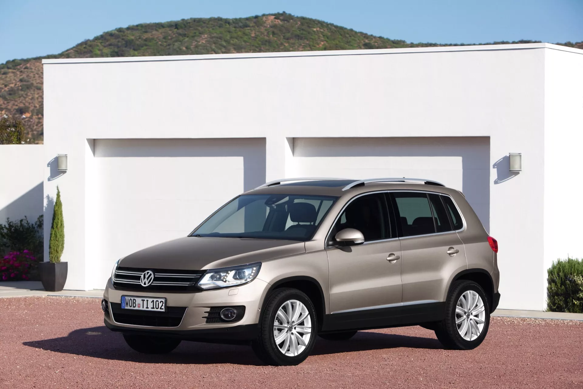 Volkswagen Tiguan z 2007 roku zaparkowany na podjeździe przed nowoczesnym domem, pokazujący jego felgi aluminiowe, charakterystyczne reflektory, światła do jazdy dziennej i relingi dachowe, na tle jasnego światła dziennego