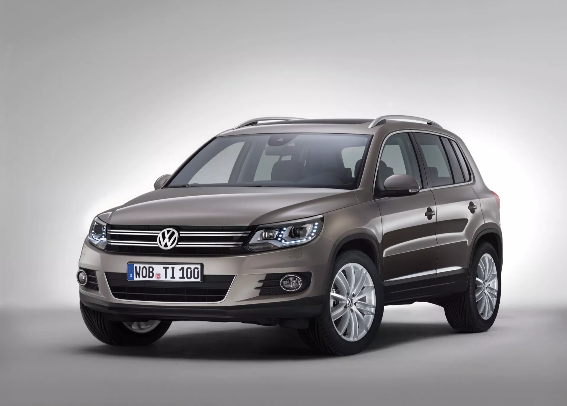 Zdjęcie studyjne Volkswagen Tiguan 2007 prezentujące lewą przednią stronę i profil boczny na jednolitym tle z delikatnym cieniem od studyjnego oświetlenia, podkreślające chromowaną atrapę, eleganckie zespoły reflektorów, światła przeciwmgielne osadzone w czarnej oprawie, felgi z lekkich stopów z wieloma szprychami, lusterka boczne w kolorze nadwozia z wbudowanymi kierunkowskazami i relingi dachowe