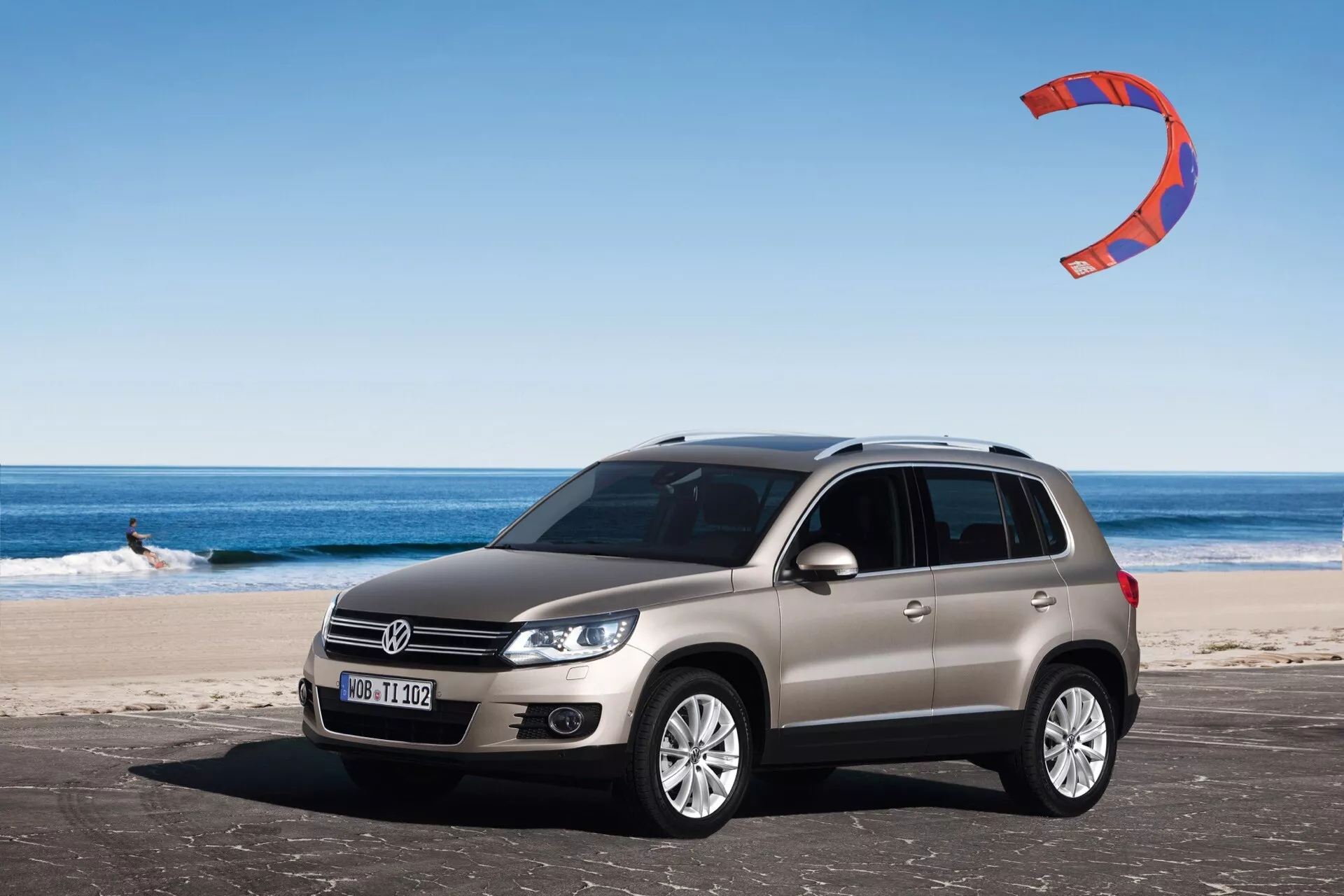 Widok z przodu i boku na srebrne SUV Volkswagen Tiguan z 2007 roku, zaparkowane na piaszczystej plaży, z widocznym logo na grillu, halogenowymi reflektorami, felgami ze stopu z wieloma szprychami, i kitesurferem w tle pod bezchmurnym niebem