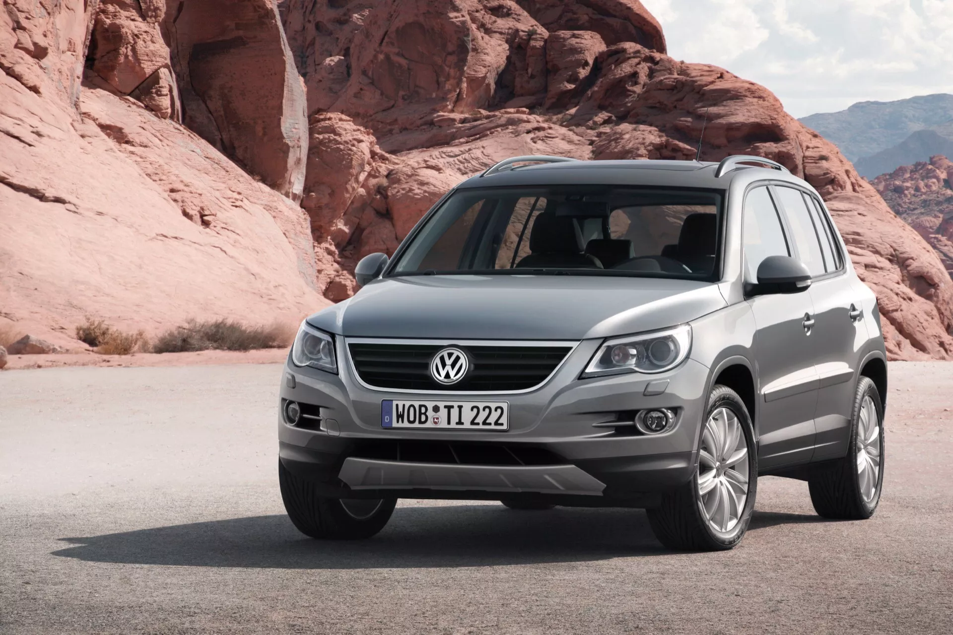 Metaliczno-szary SUV VW Tiguan z 2007 roku zaparkowany na piaszczystym terenie z dużymi formacjami skalnymi w tle, widok z przodu lewej strony pokazujący grill, reflektory, przedni zderzak z lampami przeciwmgielnymi i czujnikami, rzeźbioną maskę i felgi aluminiowe