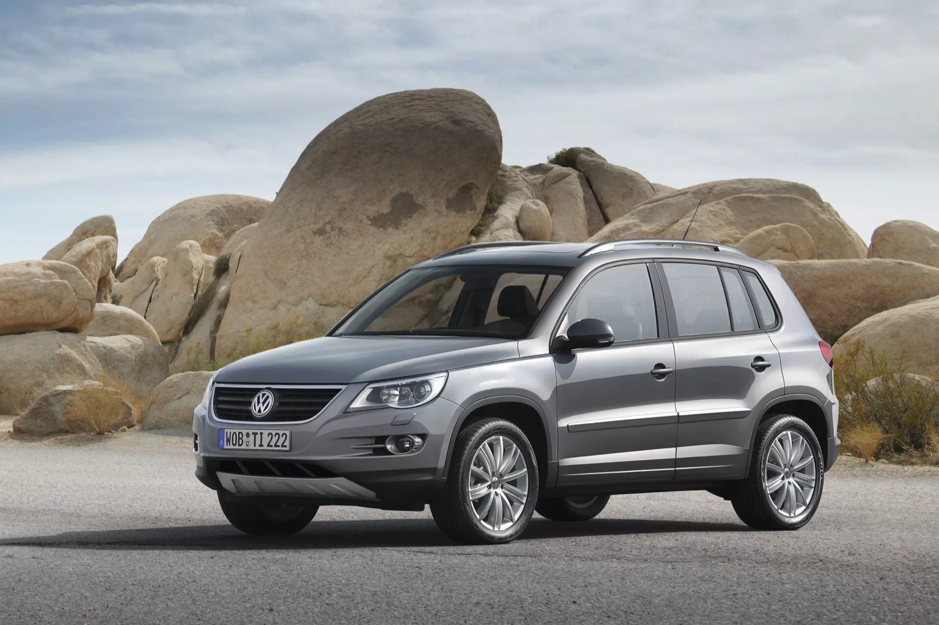 SUV Volkswagen Tiguan z 2007 roku zaparkowany na suchej, płaskiej powierzchni z dużymi zaokrąglonymi głazami w tle, prezentujący przedni i boczny profil pojazdu, w tym grill, reflektory, przedni zderzak, maskę, koła, panele boczne, drzwi i boczne okna z felgami ze stopu, relingami dachowymi i metalicznym wykończeniem lakieru.