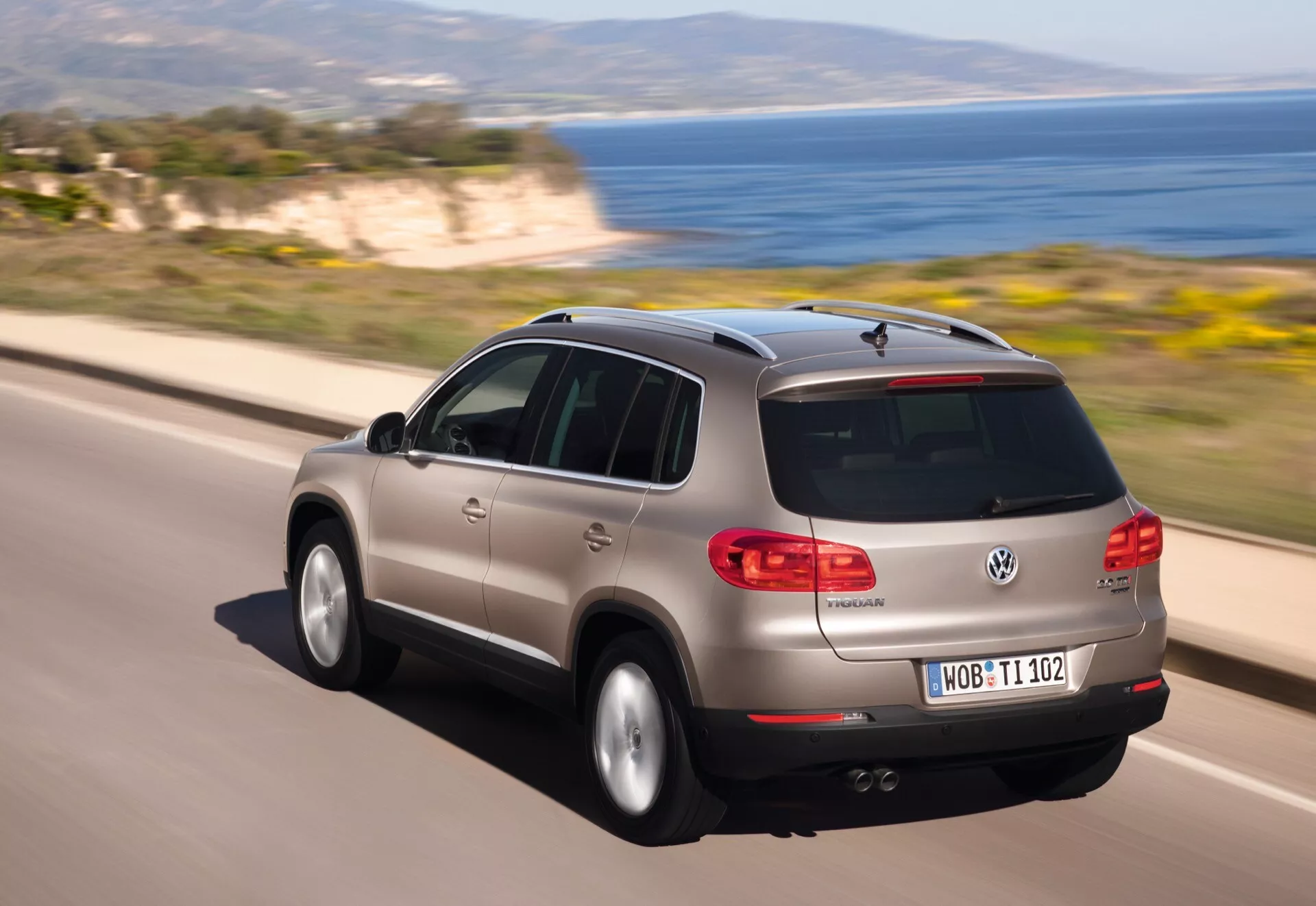 Dynamiczny tylny widok trzech czwartych poruszającego się srebrnego Volkswagena Tiguan na drodze przybrzeżnej, podkreślający jego tylny panel boczny, projekt klapy bagażnika, ledowe tylne światła oraz cechy aerodynamiczne takie jak pochylona tylna szyba, spojler dachowy oraz antena płetwy rekina.