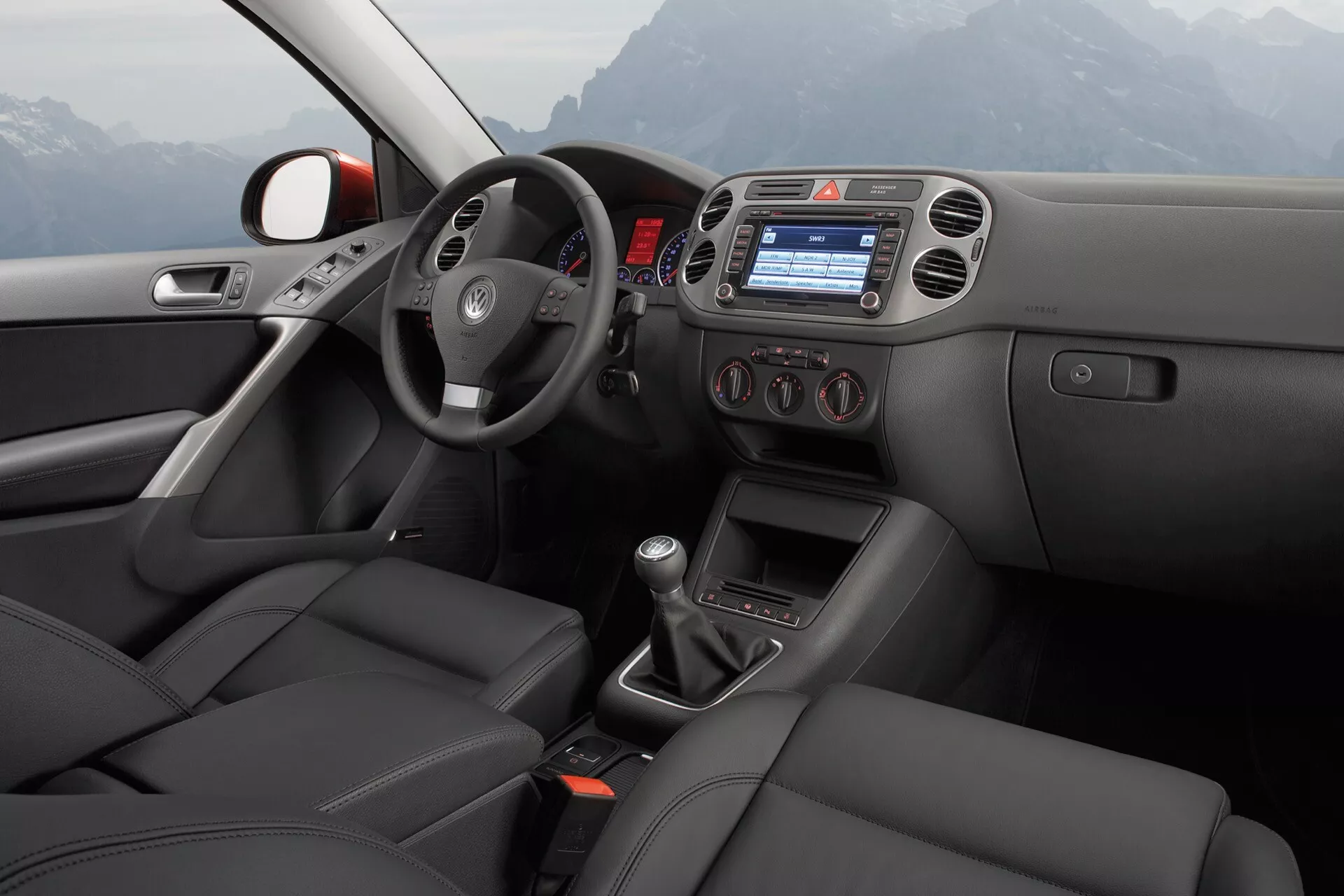 Widok wnętrza VW Tiguan 2007 z perspektywy tylnej części pasażerskiej, przedstawiający drzwi kierowcy z elektrycznymi sterowaniami okien i luster, kierownicę z zamontowanymi sterowaniami, konsolę centralną z wbudowanym wyświetlaczem systemu infotainment, kontrolę klimatyzacji, ręczną dźwignię zmiany biegów, sportowe siedzenia obite skórą, w nienagannym stanie
