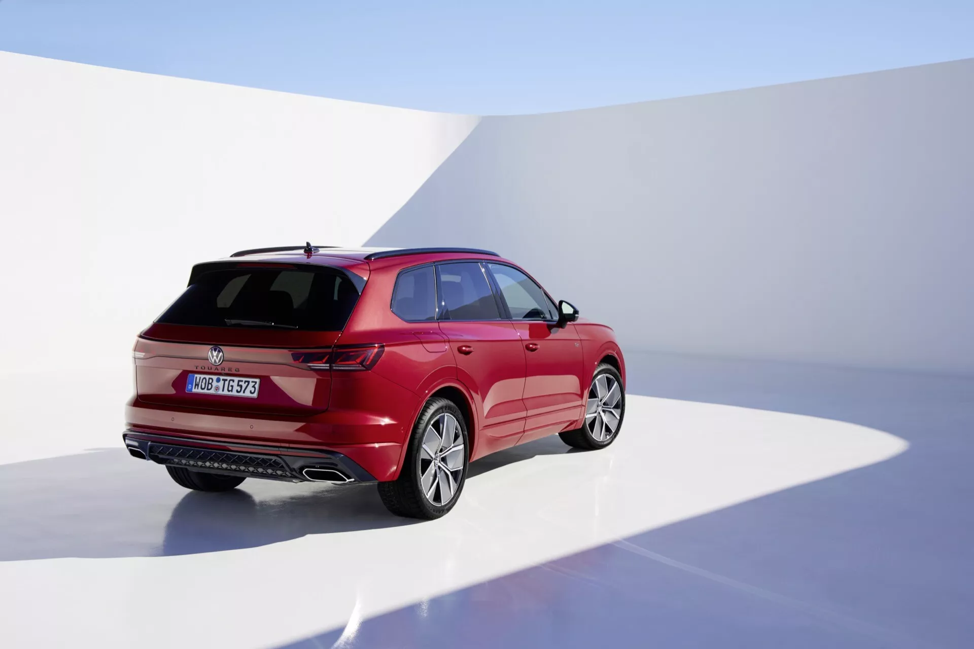 Model VW Touareg III facelift 2023 sfotografowany z tylnego ujęcia trzy czwarte podkreślającego prawą stronę, w studio z ciągłym tłem, prezentujący jego błyszczące, czerwone wykończenie, aerodynamiczny design z wyraźnymi liniami nadwozia, tylne światła LED, wycieraczkę tylnej szyby, spoiler montowany na dachu i koła ze stopu.