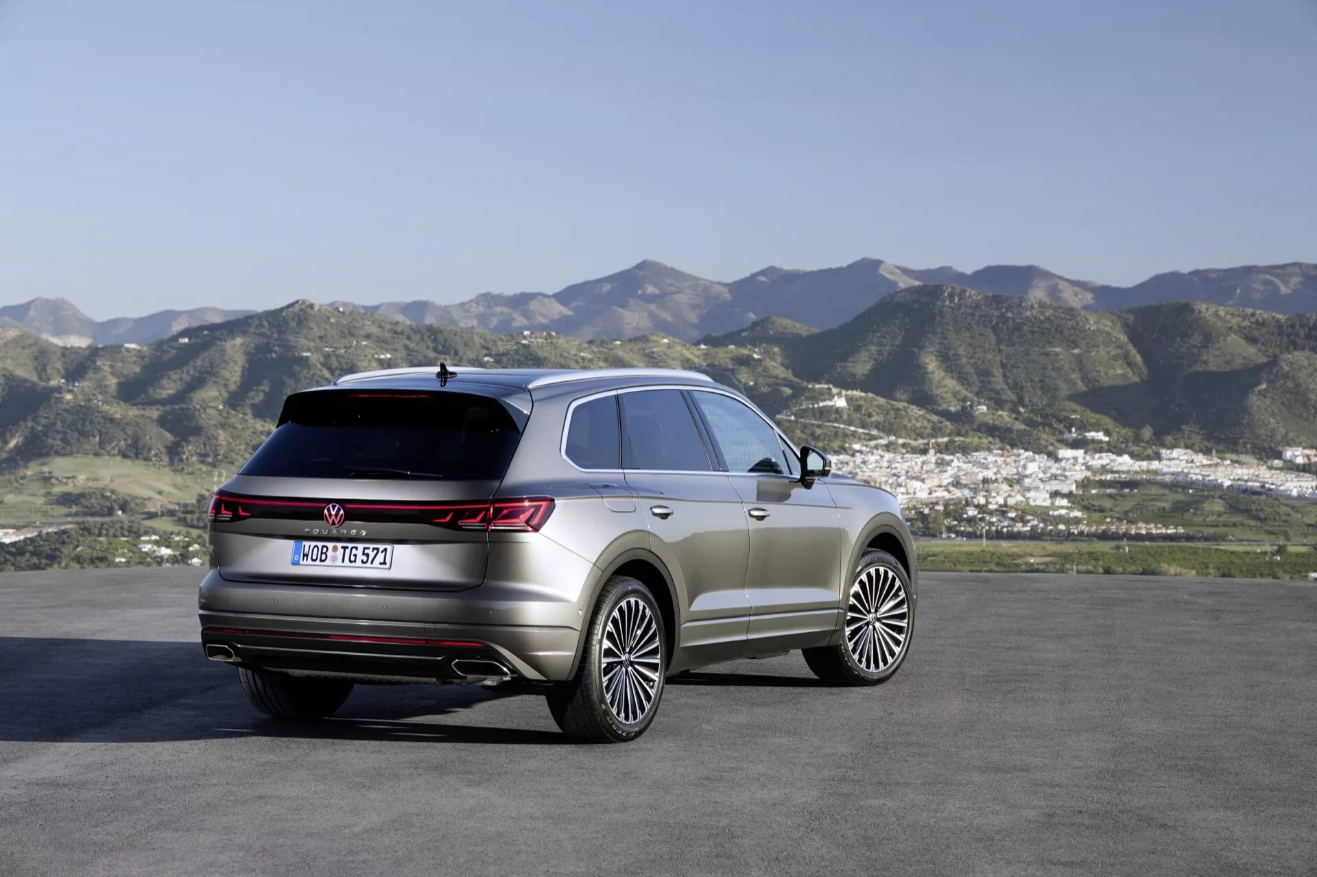SUV VW Touareg 2023 (III lifting) zaparkowany na zewnątrz z malowniczym tłem wzgórz i małego miasta lub obszaru podmiejskiego, prezentujący prawy tylny kwartał i boczny profil, z naciskiem na tylne światła, linię dachu i linie konturu nadwozia. Cechy obejmują duże koła z lekkich stopów, panoramiczny dach, tylne światła LED i premium elementy stylizacji off-road.