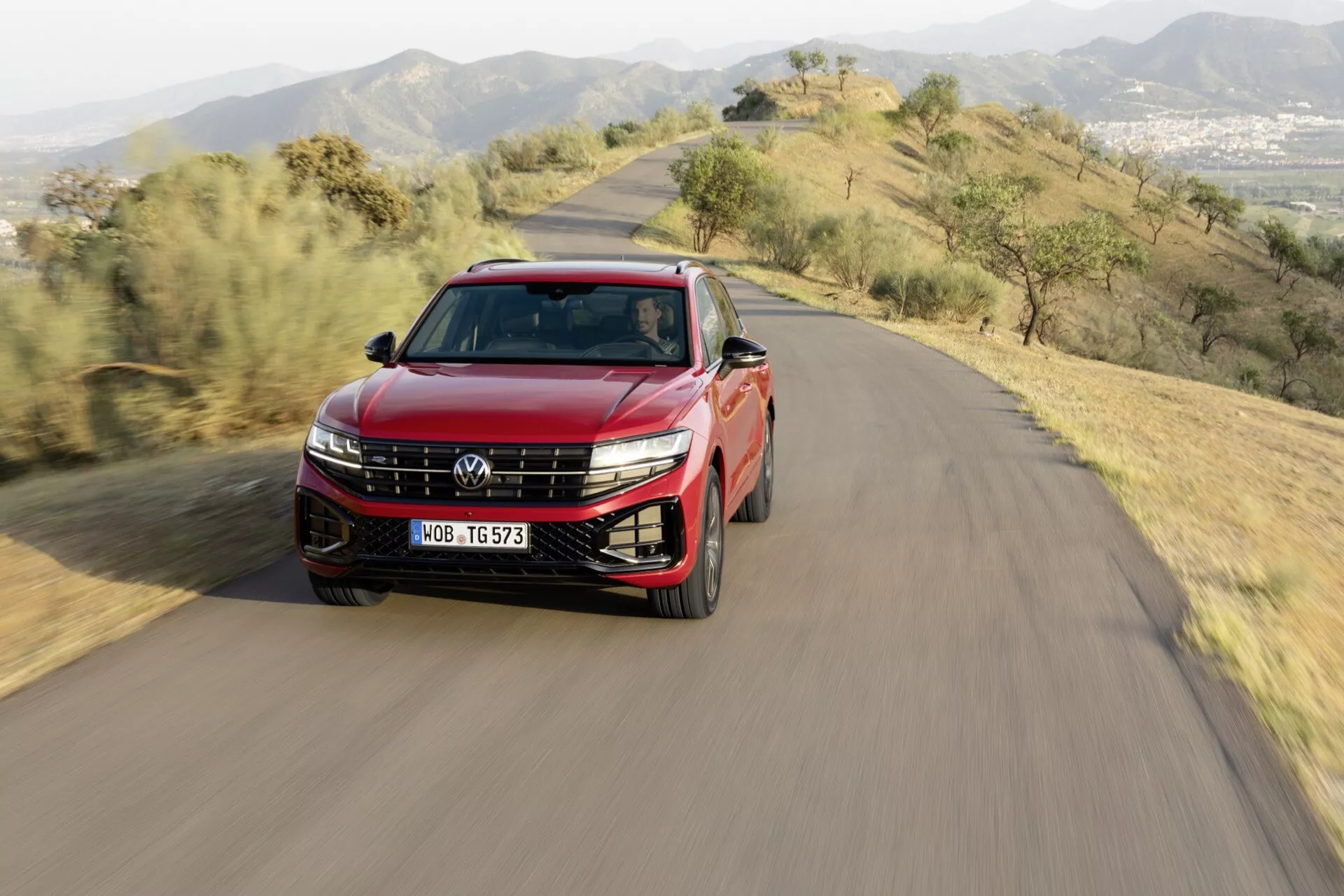VW Touareg 2023 (III Facelift) crossover/SUV jadące po krętej drodze wzgórz, z reflektorami LED, obręczami z alloy, relingami dachowymi, panoramicznym dachem, dwukolorowym lakierem i lusterkami bocznymi z kierunkowskazami, ujęcie z lotu ptaka, perspektywa frontowo-boczna w świetle dziennym