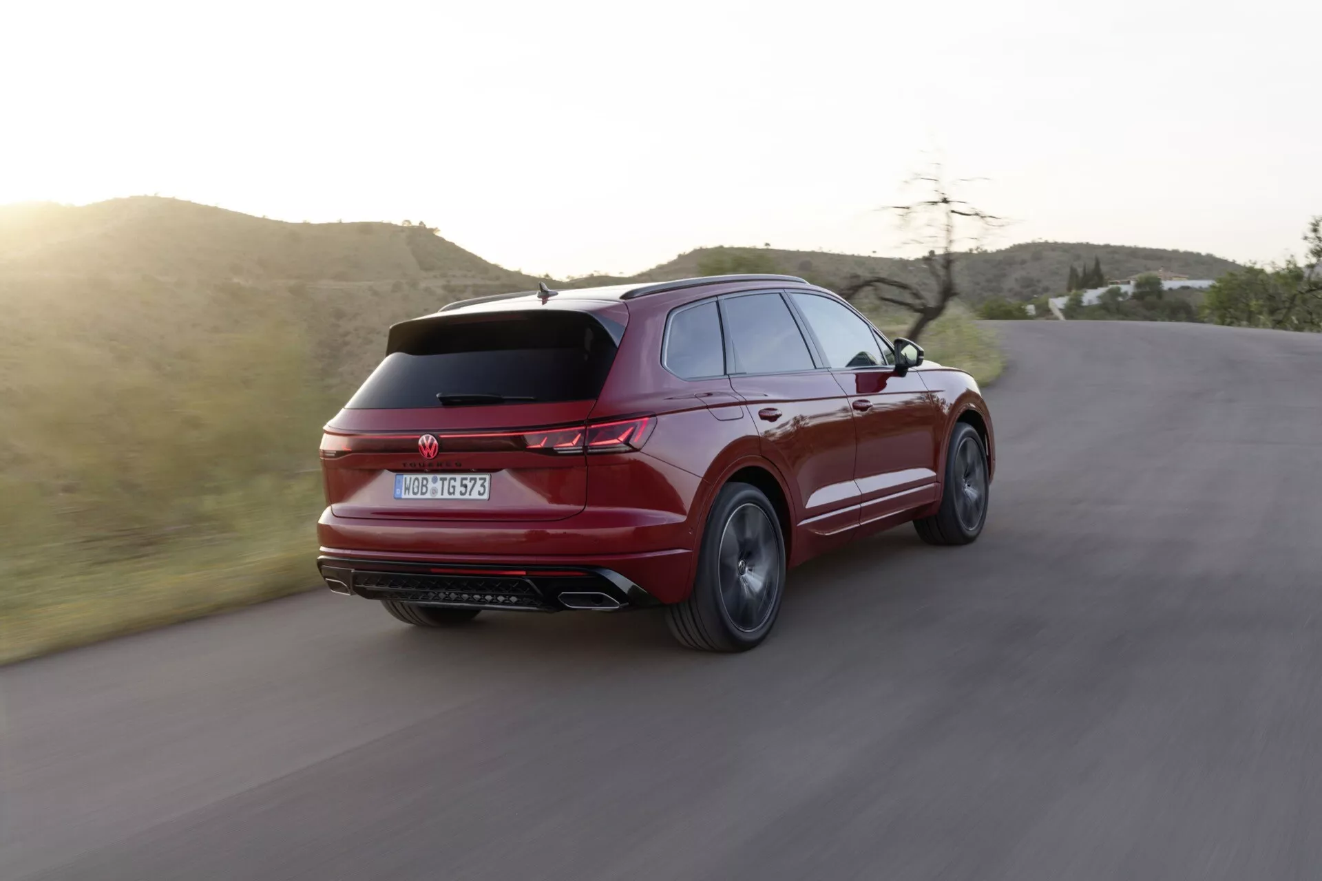Volkswagen Touareg III facelift 2023 uchwycony w dynamicznym trzykwartalnym widoku z tyłu na krętej drodze, podkreślający jego sportowy i elegancki design, smukły tylny spoiler, charakterystyczne tylne światła, podwójne rury wydechowe i wieloramienne felgi aluminiowe na tle półpustynnego krajobrazu podczas złotej godziny
