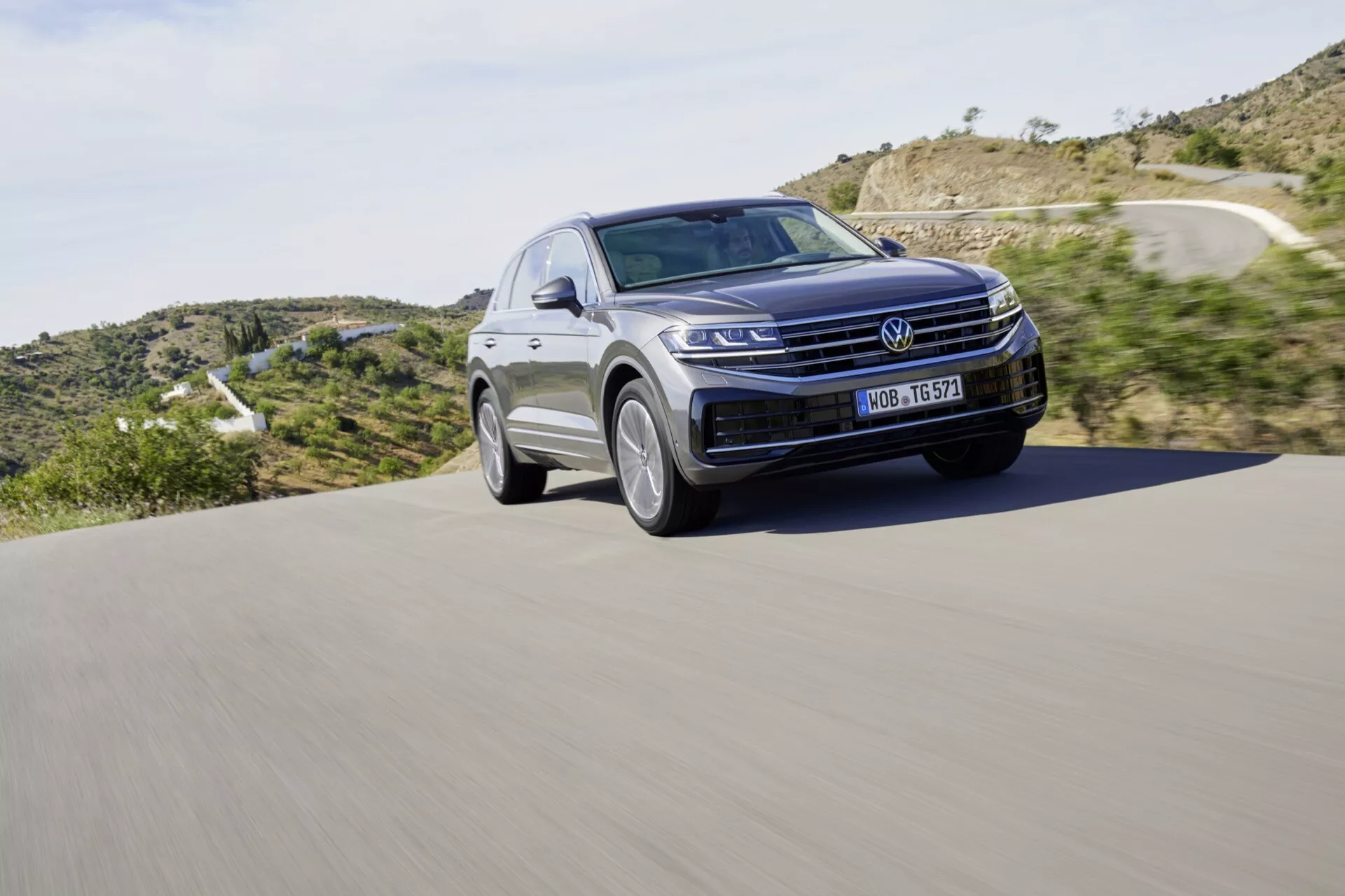 SUV Volkswagen Touareg III Facelift 2023 w ruchu na asfaltowej drodze, z wyraźną przednią kratą, światłami LED i eleganckim konturem maski na tle krajobrazu naturalnego, podkreślającym nowoczesny design i technologię motoryzacyjną.
