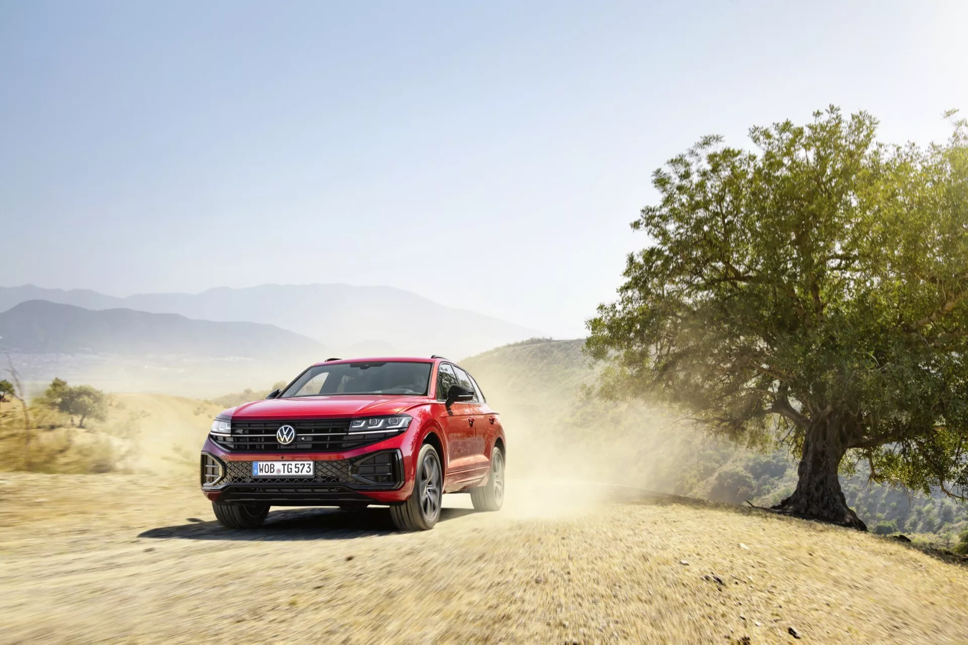 Czerwone SUV VW Touareg 2023 w dynamicznym ruchu na nieutwardzonej drodze ze śladem kurzu, prezentujące fasadę przednią, reflektory LED, grill z potencjalnymi czujnikami wspomagającymi kierowcę i felgi aluminiowe, wskazujące na terenowe zdolności i nowoczesne funkcje technologiczne