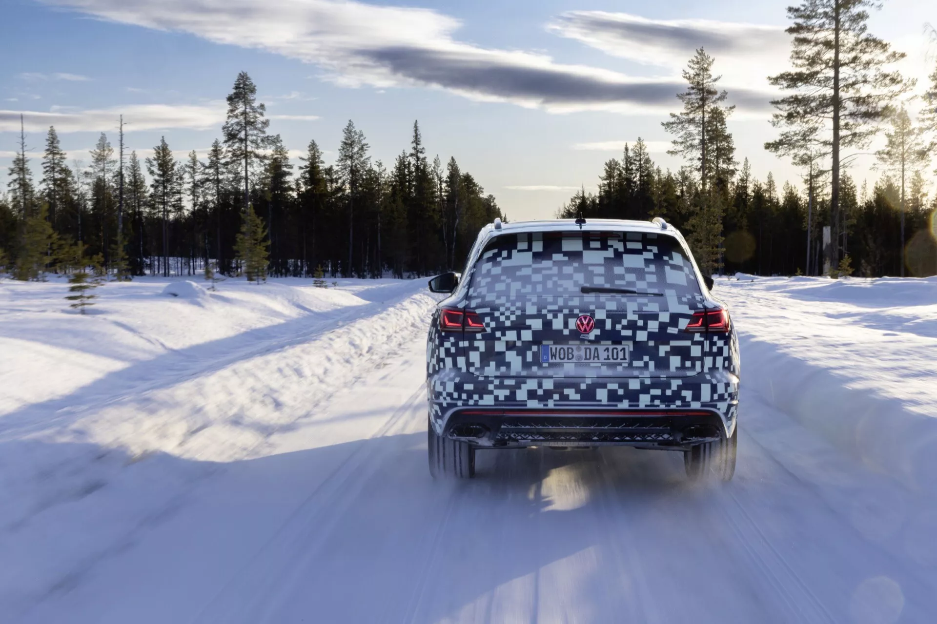 Tylny widok Volkswagen Touareg 2023 (III-Facelift) jadącego po drodze pokrytej śniegiem w zimowym lesie, pokazujący świecące tylne światła, przejrzystą tylną szybę, podwójne wydechy i spojler dachowy.