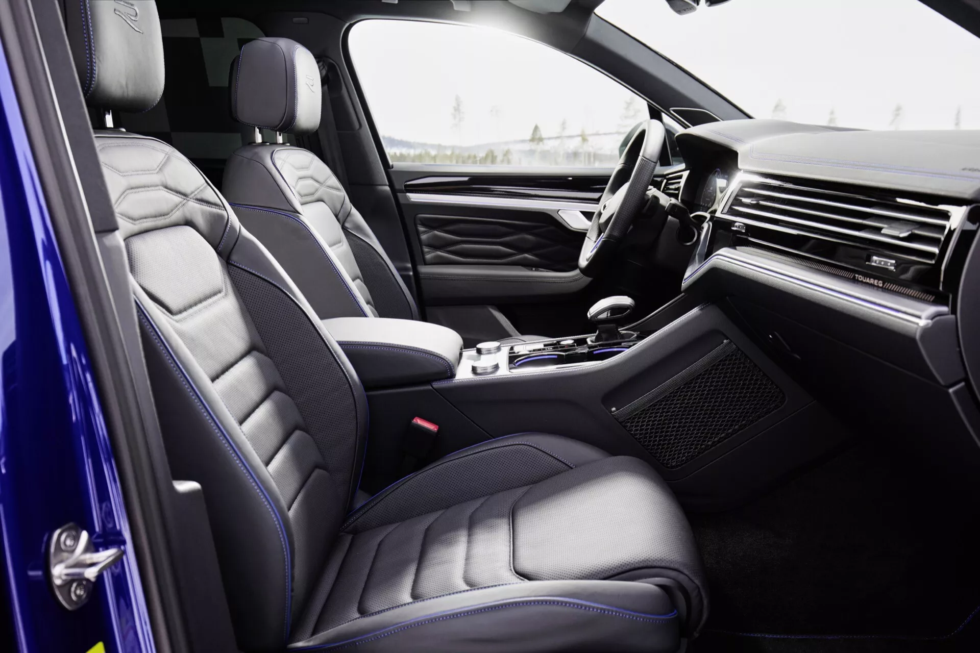 Wnętrze Volkswagen Touareg 2023 (III lifting) z perspektywy pasażera, podkreślające fotele z wysokiej jakości skóry z pikowanym wzorem, elegancka deska rozdzielcza z metalicznym lub drewnianym wykończeniem, luksusowa konsola środkowa z selektorem automatycznej skrzyni biegów, panel drzwi z wysokim poziomem opcji i częściowo widoczne koło kierownicy z zintegrowanymi kontrolkami.