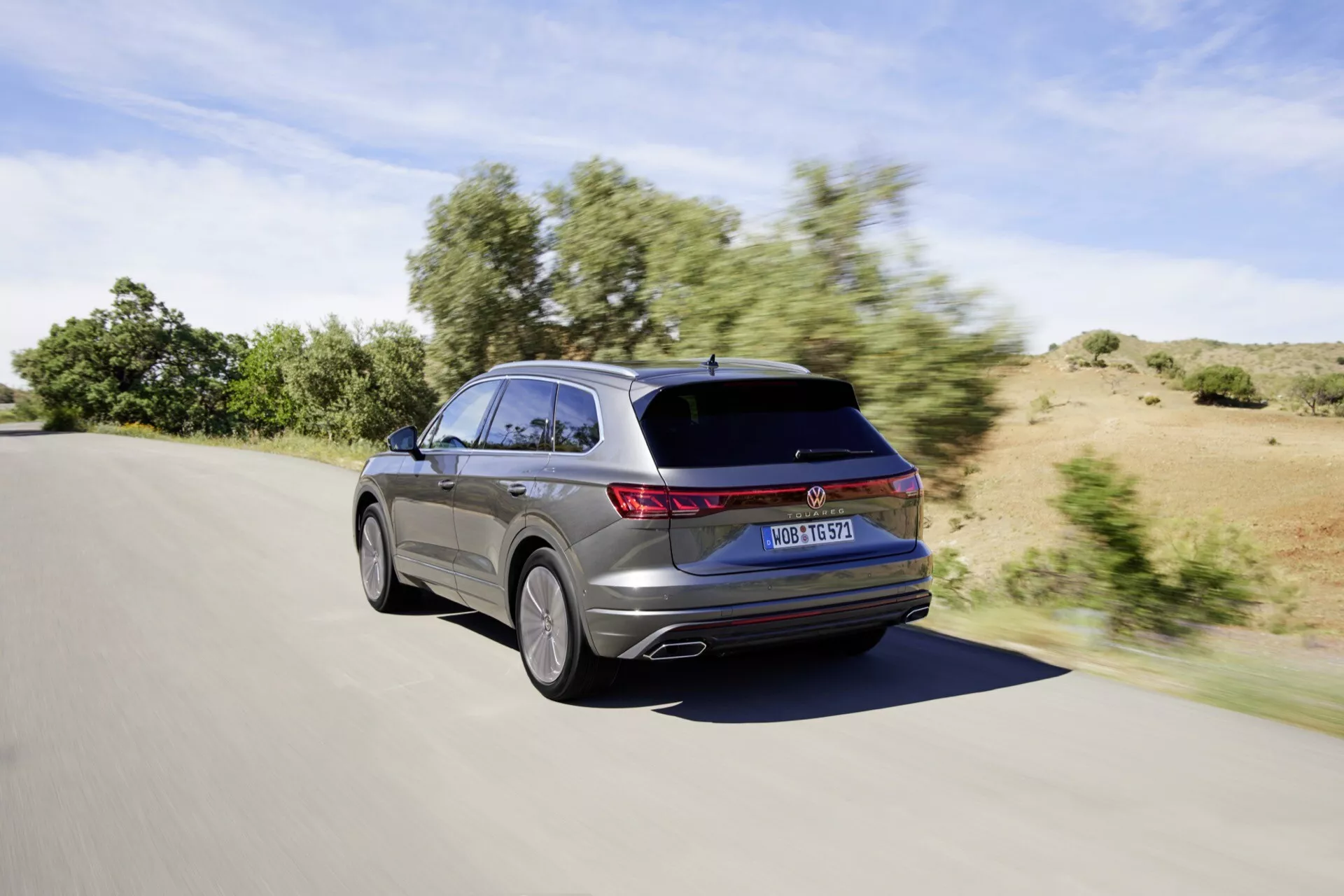 Facelifting VW Touareg III z 2023 roku ukazany z tylnego trzykwartalnego ujęcia w ruchu w ciągu dnia przy jasnej pogodzie, pokazujący LEDowe światła tylne, elegancki design kombi ze spadzistym dachem i spojlerem dachowym, częściowy widok na design kół ze stopu metali i metaliczne wykończenie lakieru.