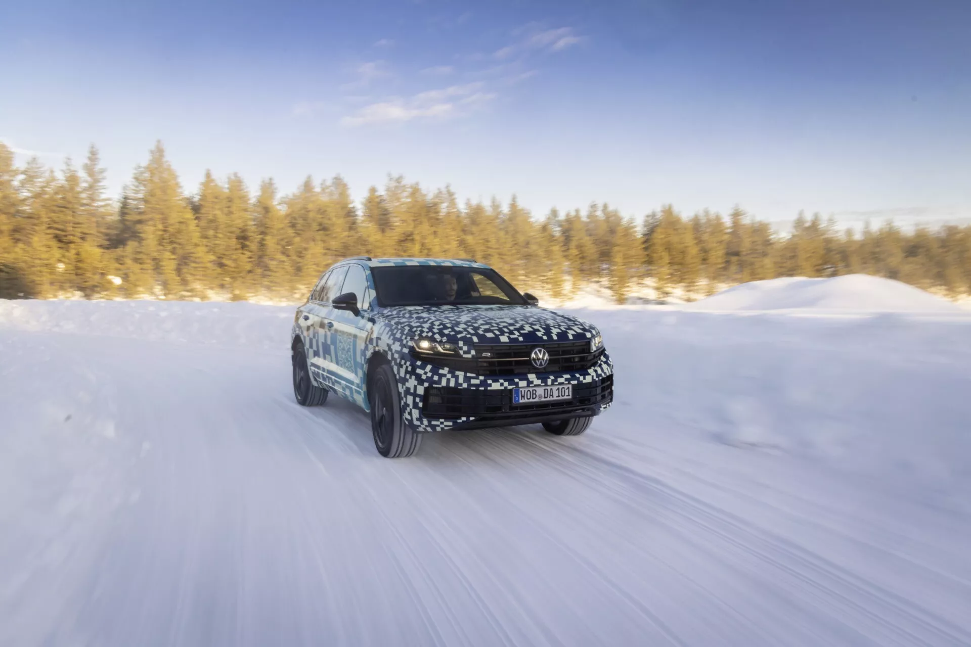 Model Volkswagen Touareg III facelift z 2023 roku dynamicznie jadący po śnieżnej drodze na słońcu, pokazujący ruch do przodu, reflektory LED, felgi aluminiowe oraz jego aerodynamiczny design z kamuflażem.