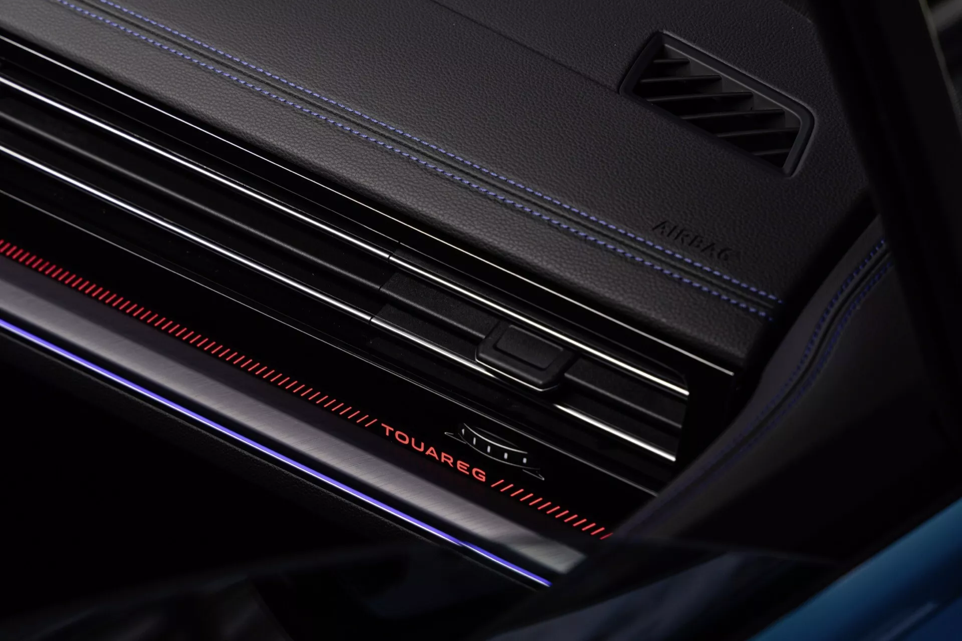 Zbliżenie na wnętrze Volkswagen Touareg po liftingu 2023 z widokiem na deski rozdzielczej, z wyraźnie widocznym napisem 'Touareg' z oświetleniem ambientowym, kratki wentylacyjne z srebrzystymi akcentami oraz atmosferyczną czerwoną listwą świetlną dla luksusowego wrażenia