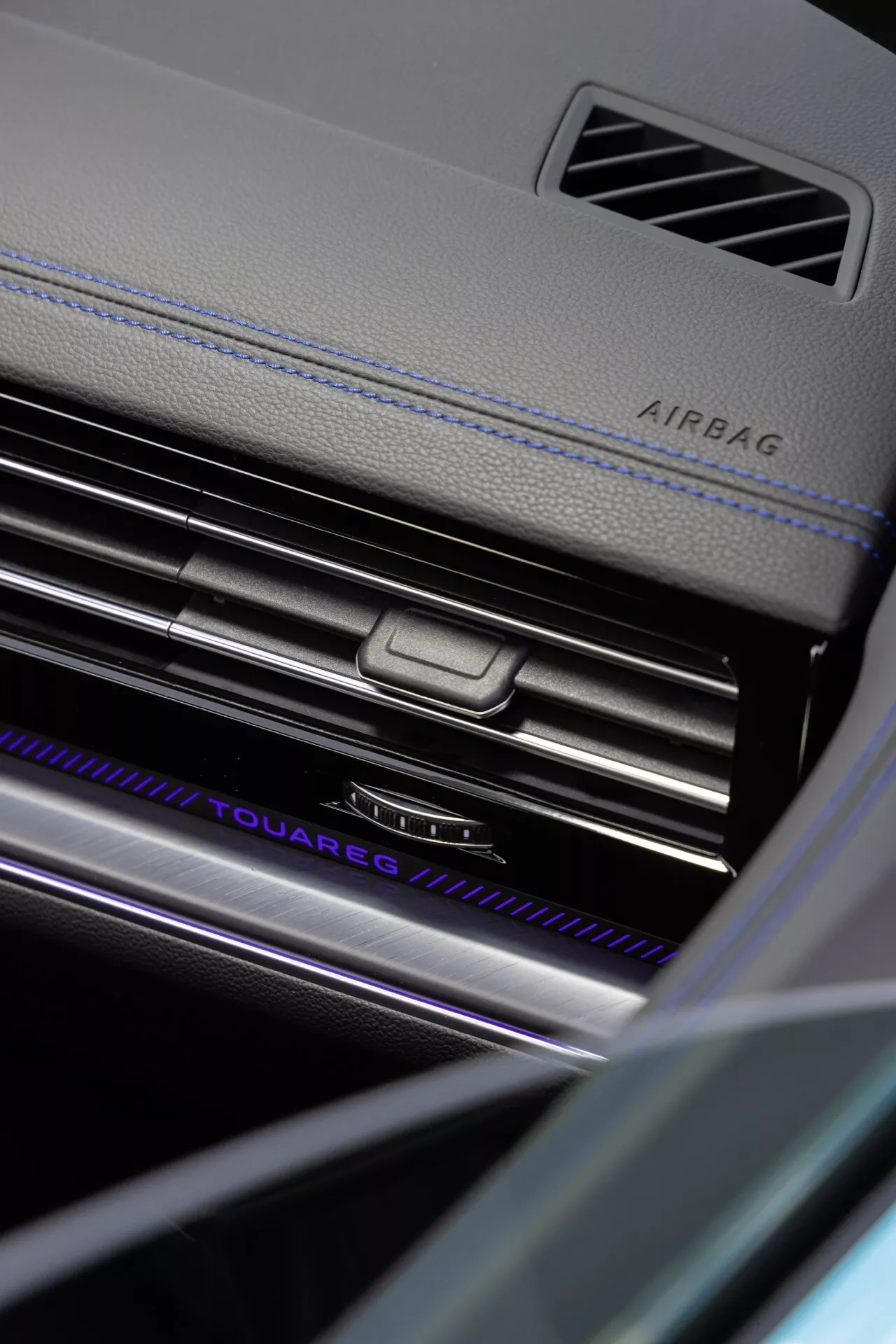 Zbliżenie na deskę rozdzielczą po stronie pasażera Volkswagen Touareg 2023 (III-Facelift) podkreślające oznaczenie AIRBAG, chromowane listwy wentylacyjne AC z centralnym pokrętłem sterującym, podświetlaną nazwę 'TOUAREG', luksusowe materiały z kontrastowym szwem oraz wyrafinowane elementy projektu wnętrza w środowisku o niskim oświetleniu, podkreślając zaawansowaną technologię oświetleniową wnętrza pojazdu i funkcje bezpieczeństwa.