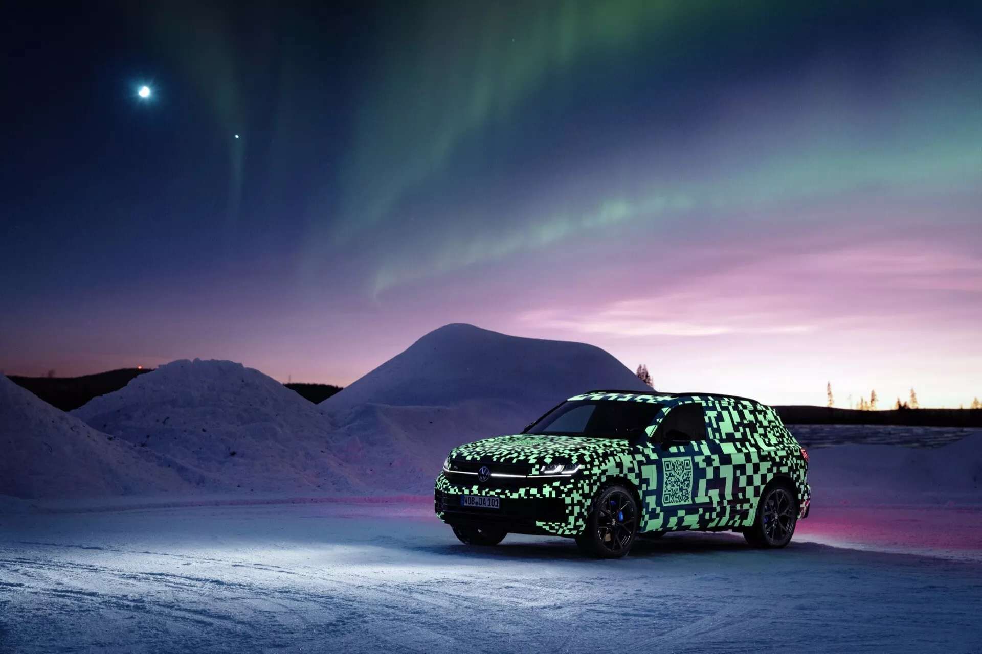 Prototyp VW Touareg (III-facelift) w kamuflażu na śnieżnej powierzchni pod zorzą polarną z jasnym ciałem niebieskim na wieczornym niebie, podkreślający zaawansowane oświetlenie i potencjalną technologię elektryczną lub hybrydową.