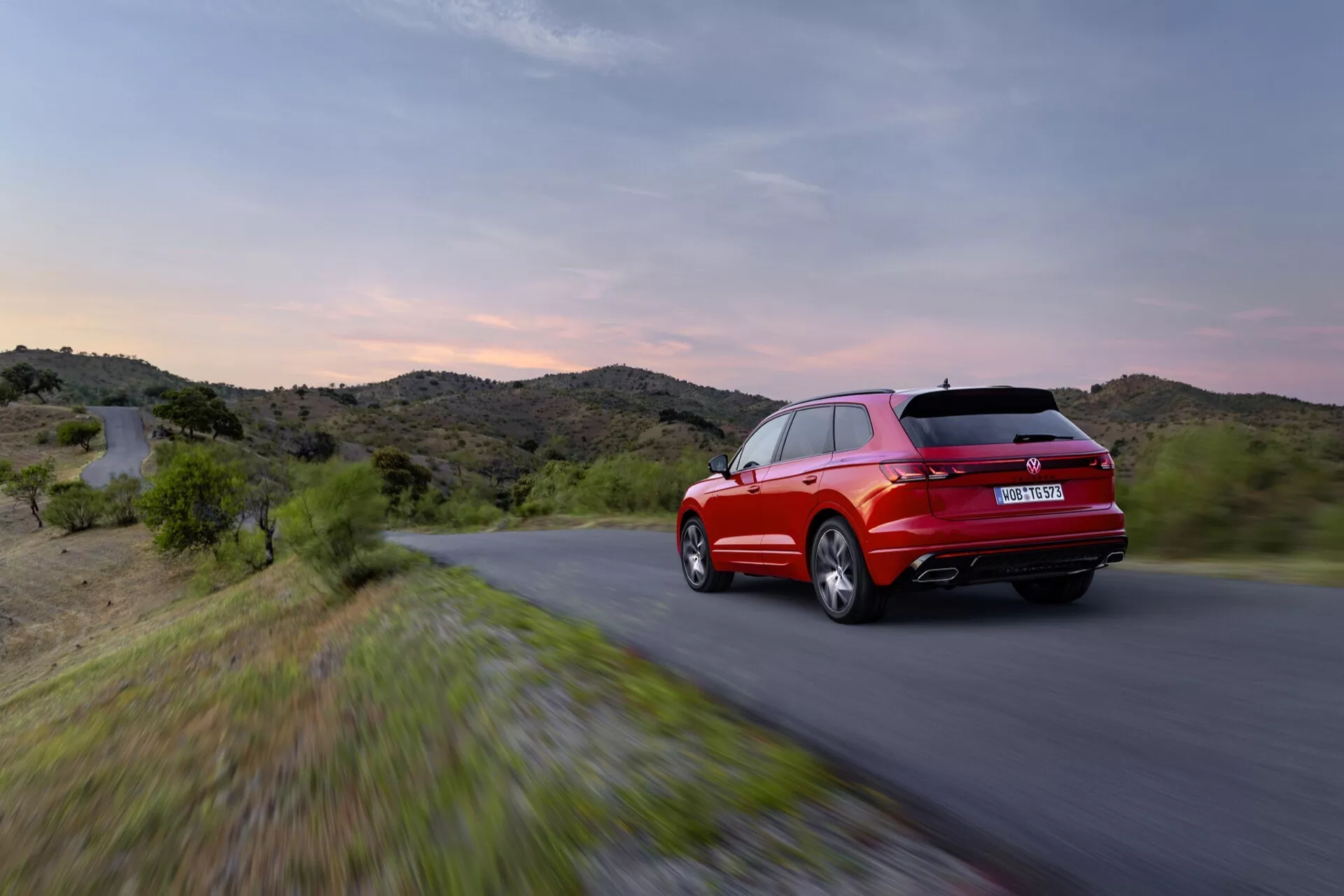 VW Touareg III Facelifting 2023 w ruchu z widoku z tyłu trzy czwarte z diodami LED i sportowym konturem na tle zmierzchu i pagórkowatego krajobrazu, podkreślającym jego czerwony kolor, aerodynamiczny design i cechy wydajnościowe.