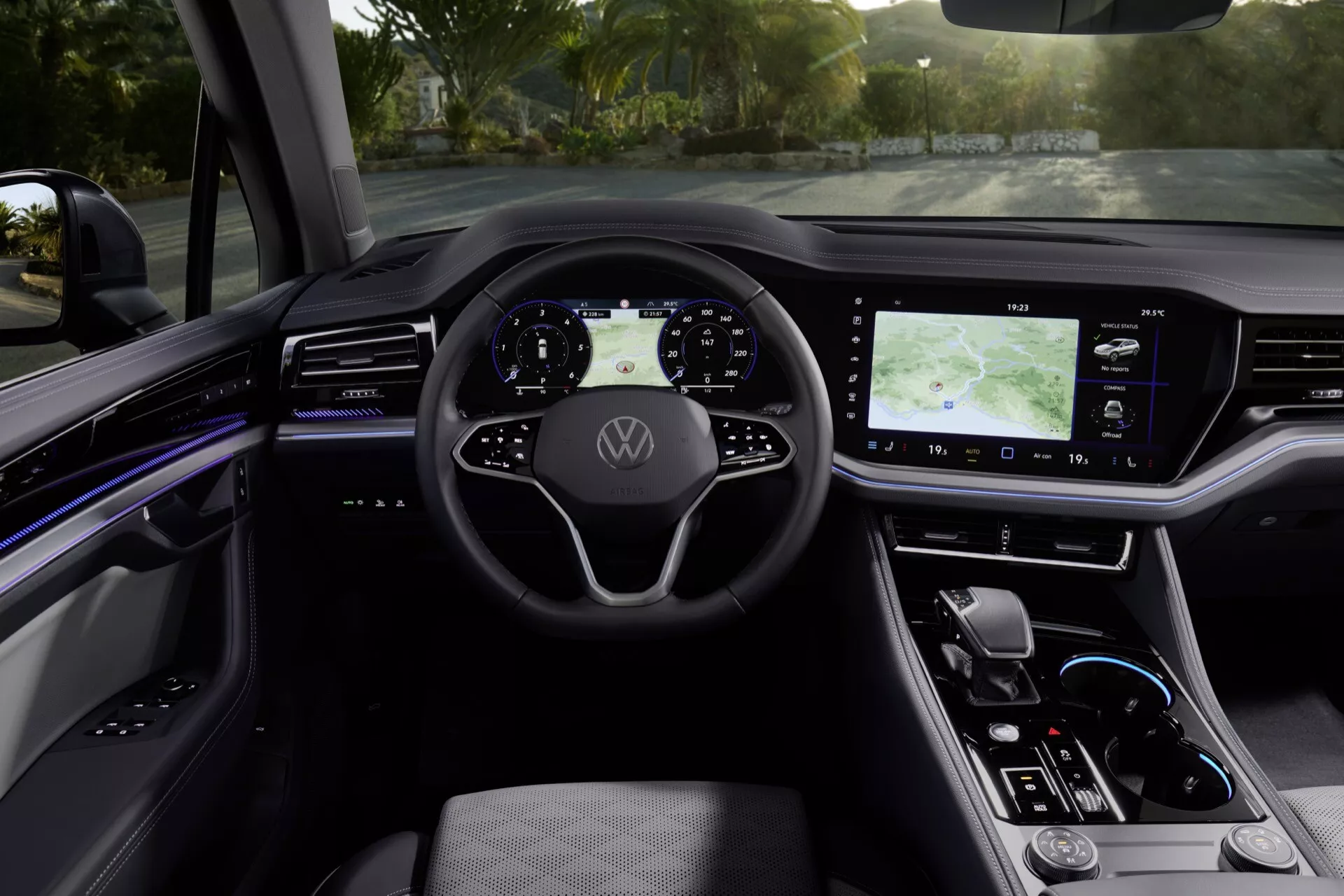 Wnętrze Volkswagen Touareg III facelift 2023 z perspektywy tylnych siedzeń w kierunku cyfrowej deski rozdzielczej oświetlonej naturalnym światłem, prezentujące wielofunkcyjną kierownicę z logo, cyfrowy zestaw wskaźników, duży system infotainment z ekranem dotykowym i GPS, eleganckie dotykowe sterowanie klimatyzacją, minimalistyczną dźwignię automatycznej skrzyni biegów oraz centralną konsolę z oświetleniem ambientowym, wysokiej jakości wykończeniem i funkcjami bezpieczeństwa