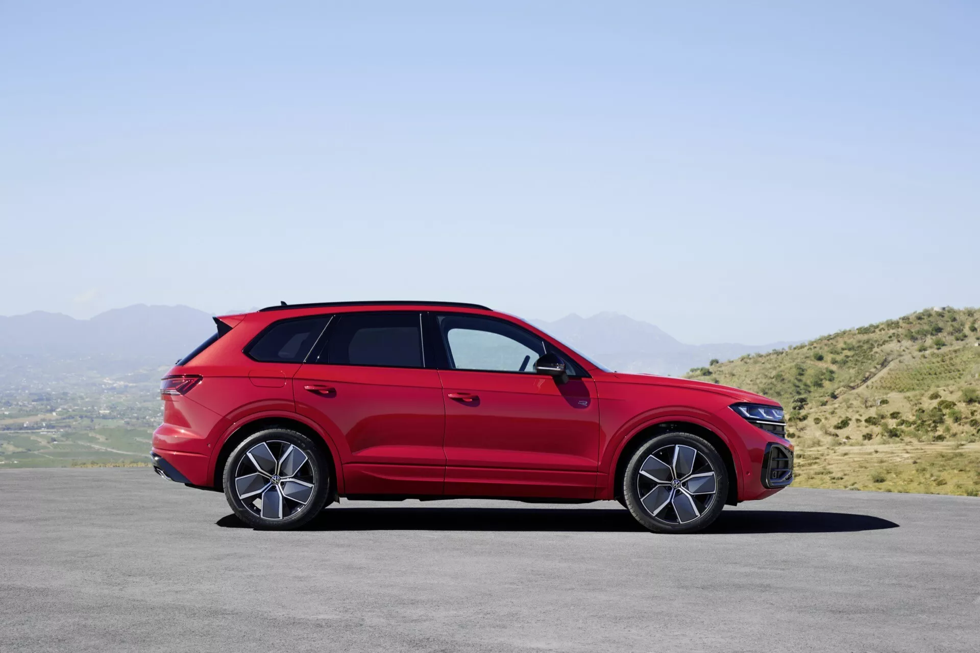 Model VW Touareg III po liftingu w kolorze czerwonym zaparkowany na płaskiej nawierzchni z krajobrazem górskim i czystym niebem w tle, prezentujący swój profil boczny z dokładnym widokiem na koła z lekkich stopów, klamki drzwi, lusterka boczne z wbudowanymi kierunkowskazami, relingi dachowe, sportową sylwetkę stylu coupe oraz przyciemniane szyby.