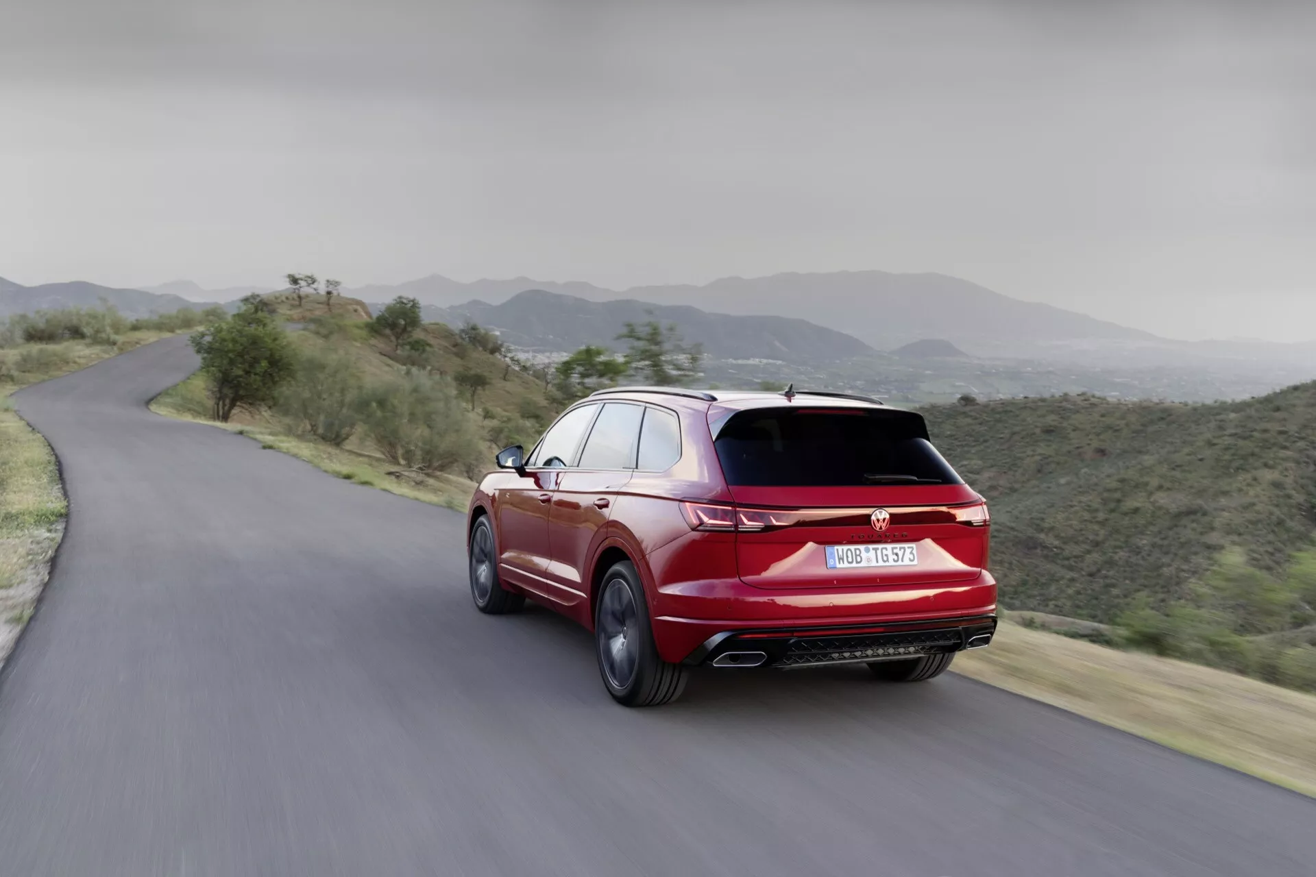 Dynamiczny tylny widok VW Touareg 2023 na górskiej drodze, prezentujący aerodynamiczny design, podwójne wydechy i sportową wersję w jaskrawoczerwonym kolorze